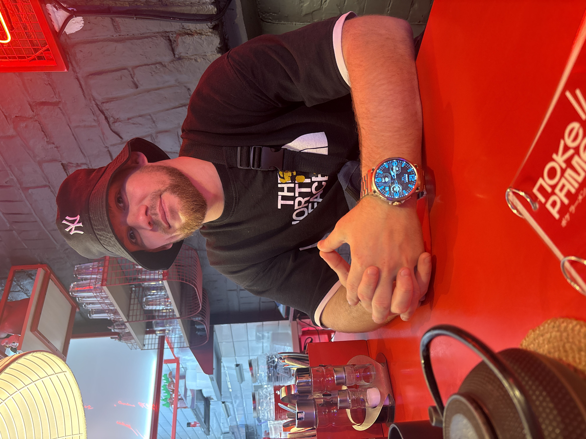 The image depicts a fair-skinned male with a beard, wearing a black North Face T-shirt and a black bucket hat with a white "NY" logo, seated at a red table. He is wearing a large, digital watch on his left wrist. Part of a "POKE // PRIME" advertisement is visible at the edge of the table. The background features a brick wall with red neon lighting, a display rack with glassware, and kitchen equipment, potentially in a restaurant setting. The location is likely in Perm, Russia, given the Russian signage. The man appears to be posing for the photo. A black teapot is on the table.