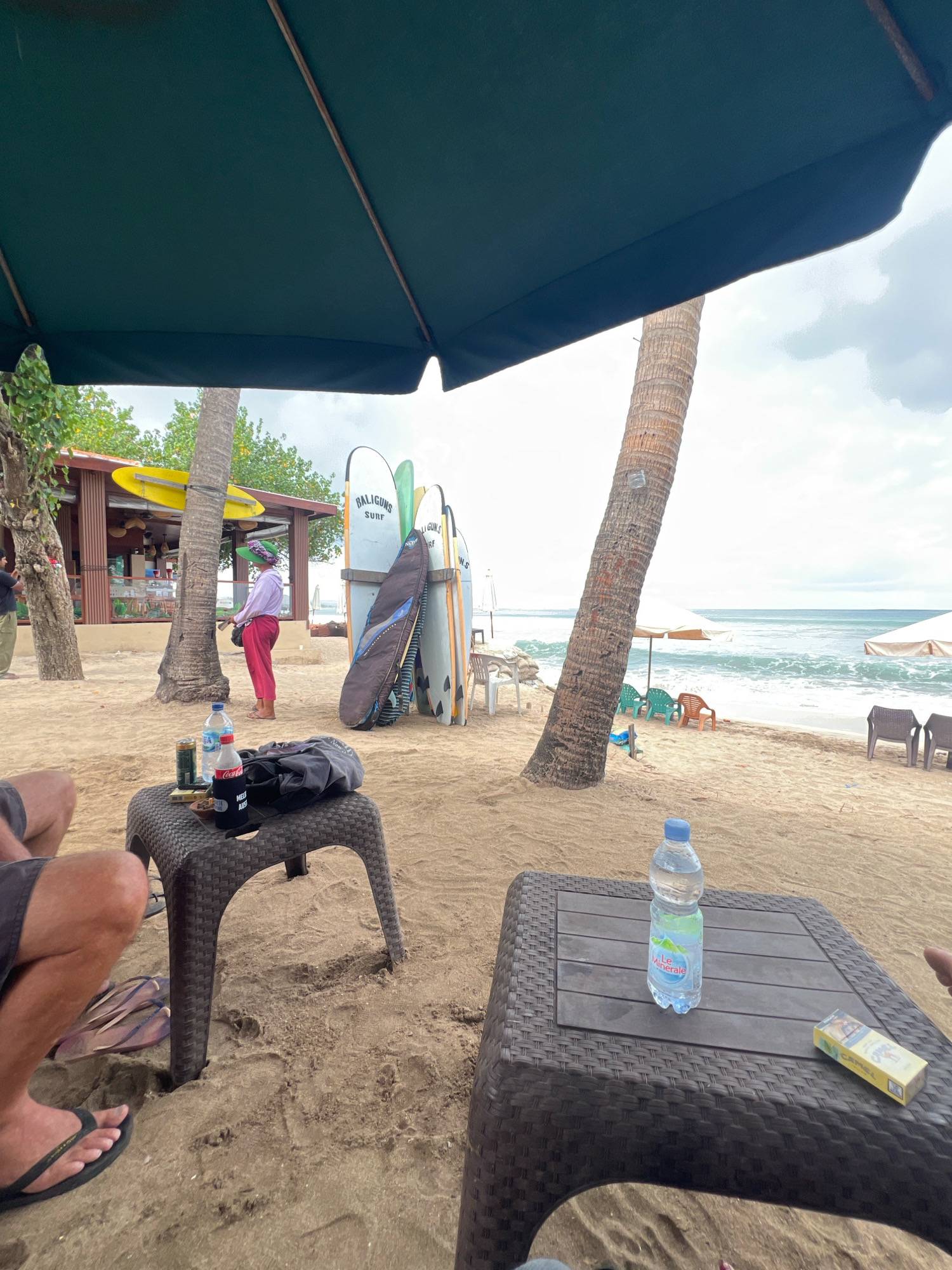This image depicts a beach scene in Kuta, Indonesia. In the foreground, two people's legs are visible, suggesting they are seated and relaxing. On a dark brown plastic table in the left foreground, there are two cans, a bottle of Coca-Cola, a bottle of water, and a black cloth bag. Another dark brown plastic table in the right foreground holds a bottle of Le Minerale water and a pack of Camel cigarettes. In the middle ground, a woman in a green hat and pink pants stands near a small building with a yellow surfboard on its roof. Several surfboards are leaning against a palm tree. The beach is sandy, with the ocean and waves visible in the background. The sky is overcast.