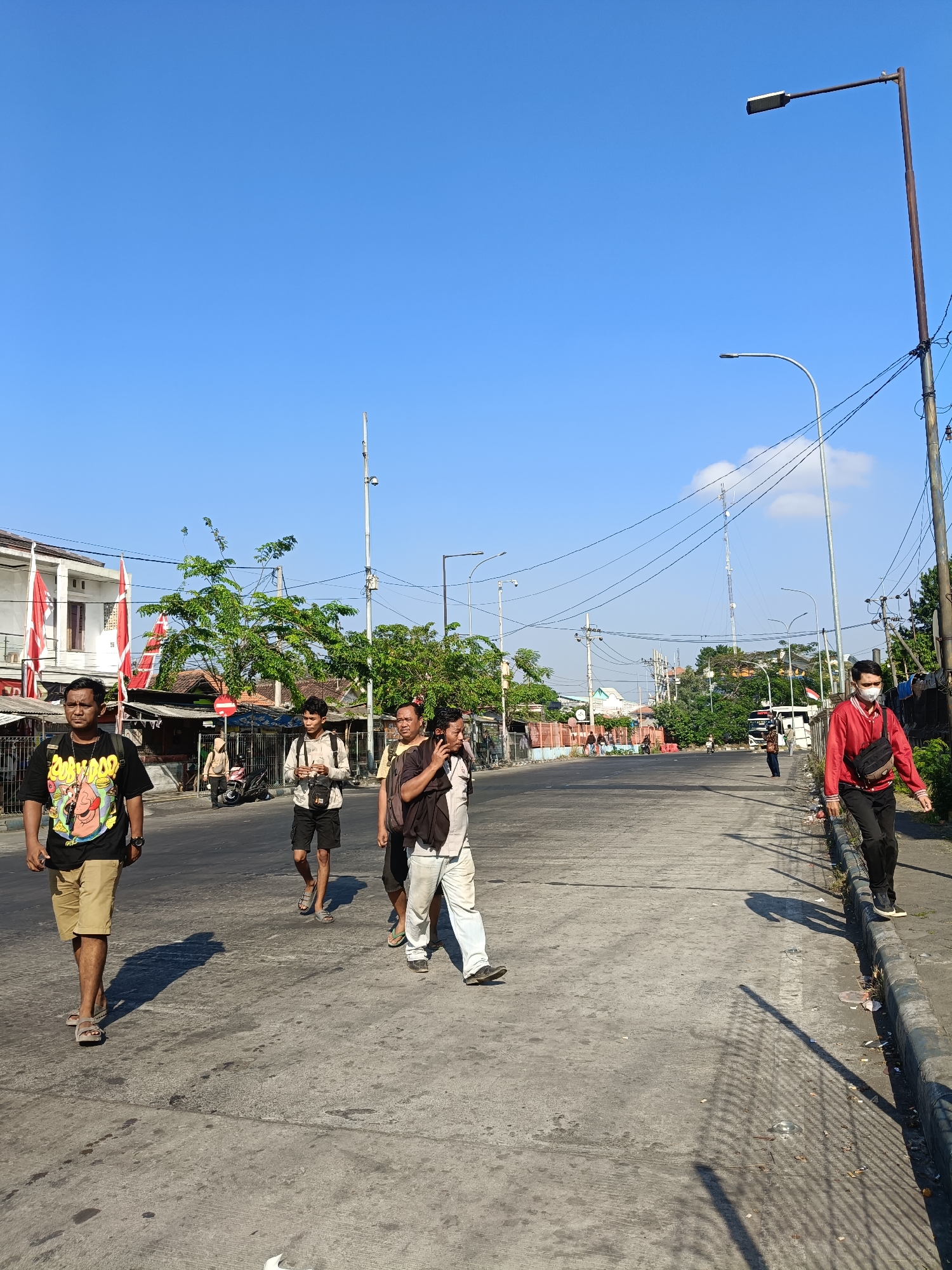 The image depicts a bright, sunny day in Kedungrejo, Indonesia. The scene is an outdoor street with a clear blue sky. Several people are visible on the asphalt road. In the foreground on the left, a man wearing a black t-shirt with a graphic print and khaki shorts walks towards the camera. To his right, a young man in a light-colored hooded jacket and dark shorts is also walking, holding a phone. Further back, another man in a yellow shirt is walking, and a person in white pants and a dark shirt is walking with them. On the right side of the road, a person in a red jacket and black pants walks away from the camera, wearing a white face mask and carrying a black bag. The street is lined with buildings and trees, and there are several utility poles with wires stretching across the scene. Red and white flags are displayed on poles in front of a building on the left. The road has visible markings and some debris along the curb.