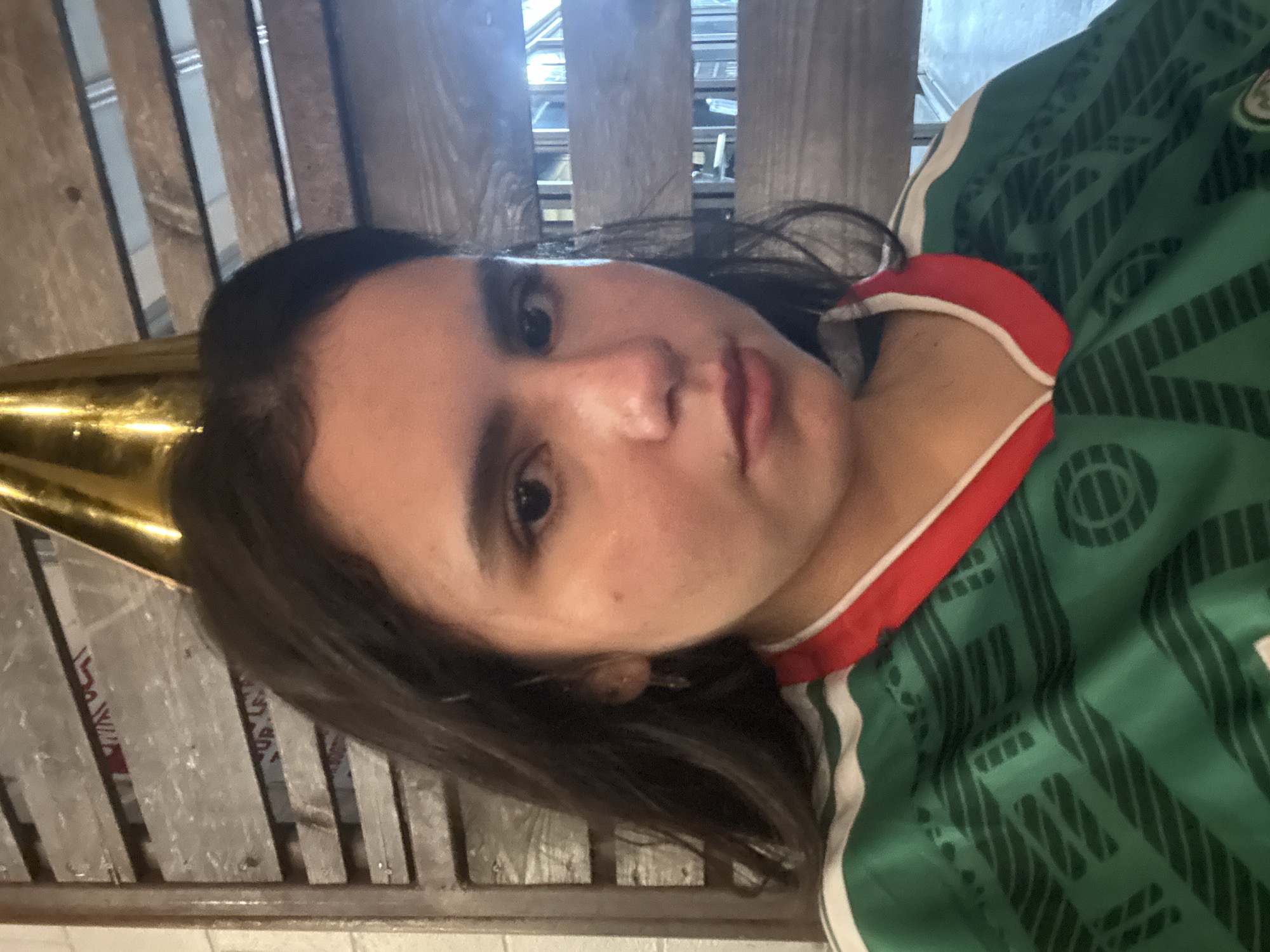 A medium shot shows a young person wearing a green jersey with red and white trim and a gold party hat. The person's face is turned slightly towards the camera, with dark hair framing their face. The background consists of weathered wooden slats, creating a rustic aesthetic. The lighting is somewhat dim, with some ambient light sources visible in the background, suggesting an indoor setting. The scene appears to be a casual social gathering or a personal moment captured for remembrance. No specific location in Austin, United States of America can be identified.
