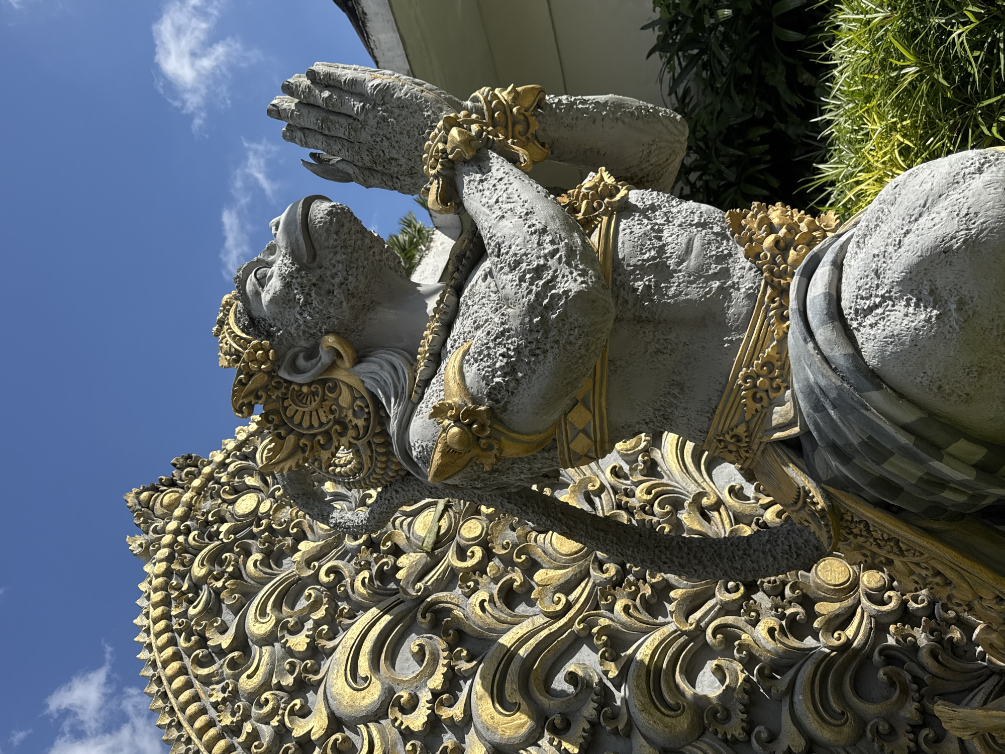 In Tegallalang, Indonesia, a stone statue with a monkey-like face and intricate golden headdress and adornments stands in prayer. The statue is depicted with hands clasped together in front of its chest, and a detailed, ornate golden structure rises behind it, featuring swirling patterns and textured elements. The statue is made of textured grey stone with a rough surface. It is adorned with golden jewelry, including bracelets on its forearms and a decorative band around its neck. A sash draped over its shoulders is also embellished with golden details. Behind the statue, lush green foliage and grass are visible, suggesting an outdoor setting. The sky above is a clear blue with scattered white clouds.