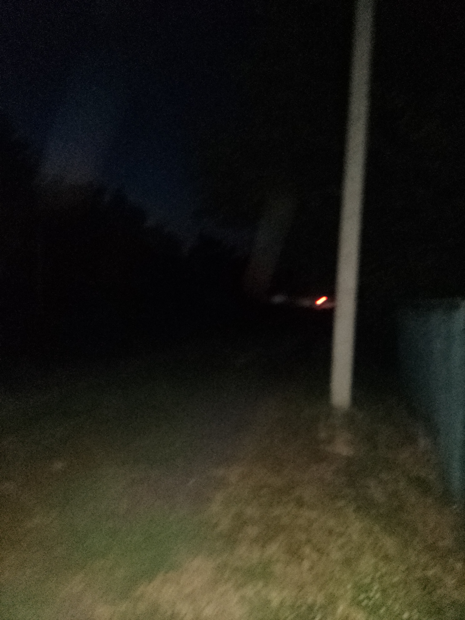 The image depicts a dark, outdoor scene at night, with poor lighting conditions. A thin, vertical, light-colored pole is present in the right half of the frame. In the background, a faint reddish-orange light source is visible. The ground appears to be covered in grass. The overall environment suggests a rural or suburban setting. The specific location in Ustyvytsia, Ukraine is not discernible from the visual information provided in the image.