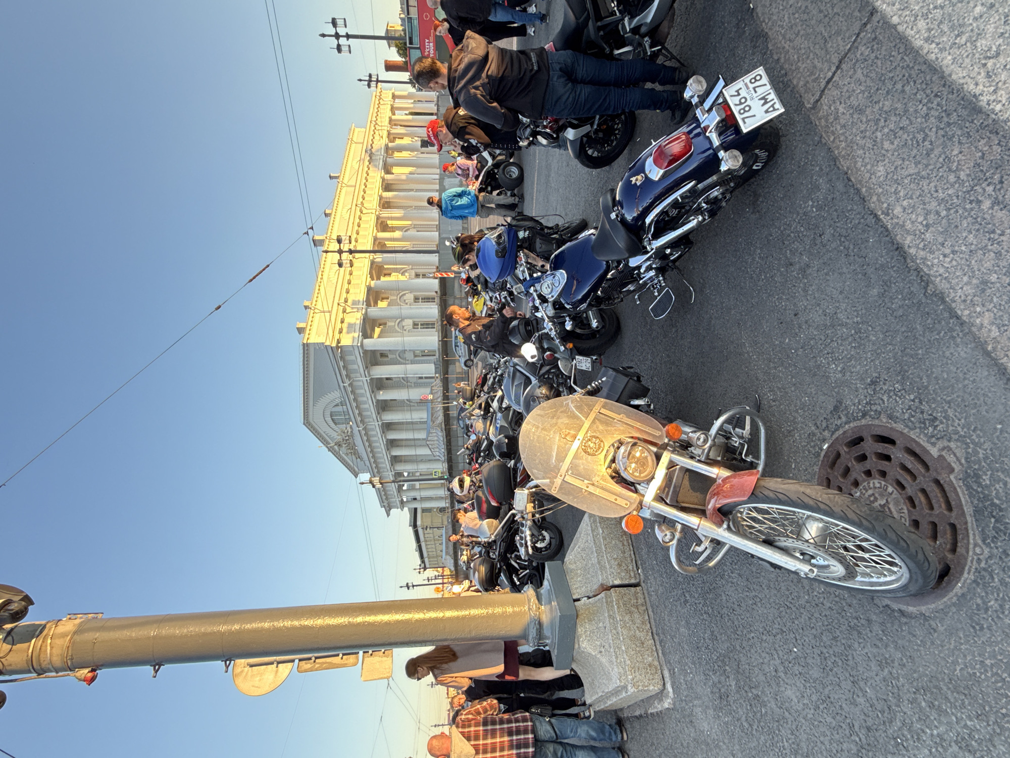 The image depicts an outdoor urban scene in Saint Petersburg, Russia, during what appears to be the golden hour due to the warm, low-angle sunlight. A large gathering of motorcycles and people occupies the paved foreground and midground, with a prominent neoclassical building in the background.
In the immediate foreground, a red and chrome cruiser-style motorcycle with a large clear windshield is parked on the left, its front wheel partially covering a round metal manhole cover. To its right, the rear of a dark blue cruiser-style motorcycle is visible, displaying a white Russian license plate with the registration "7864 RUS AM 78" (where '78' is the region code for Saint Petersburg). The ground is paved asphalt with a grey stone curb extending from the right.
A tall, cylindrical grey street pole with a textured surface stands prominently on the left, featuring multiple attached traffic signs and a small red indicator light. Its base is constructed from two rough-hewn stone blocks.
The midground is densely populated with various motorcycles and individuals. Numerous motorcycles of different types, including cruisers and sport bikes, are parked in close proximity. Many have luggage attached, such as saddlebags or top cases. A blue motorcycle helmet rests on the seat of one of the bikes. Approximately 20-30 adults are visible, scattered among the motorcycles, standing in small groups, conversing, or observing the bikes. They are dressed in casual attire. Notable figures include a man in a plaid shirt and jeans on the far left, a woman with long brown hair in a beige top and dark skirt near the street pole, and a man in a black jacket and jeans standing with his back to the camera on the right. An all-terrain vehicle (ATV) is partially visible in the background, near a person in a blue hoodie.
The background is dominated by a grand, multi-storied white neoclassical building with a long facade featuring numerous columns and pilasters, illuminated by direct, warm sunlight. This is the General Staff Building, situated on Palace Square. Several ornate black lampposts are positioned in front of the building. On the far right, a red double-decker "CITY TOUR" bus is partially visible. The sky above is clear and light blue, with a few overhead utility wires visible stretching across the frame.