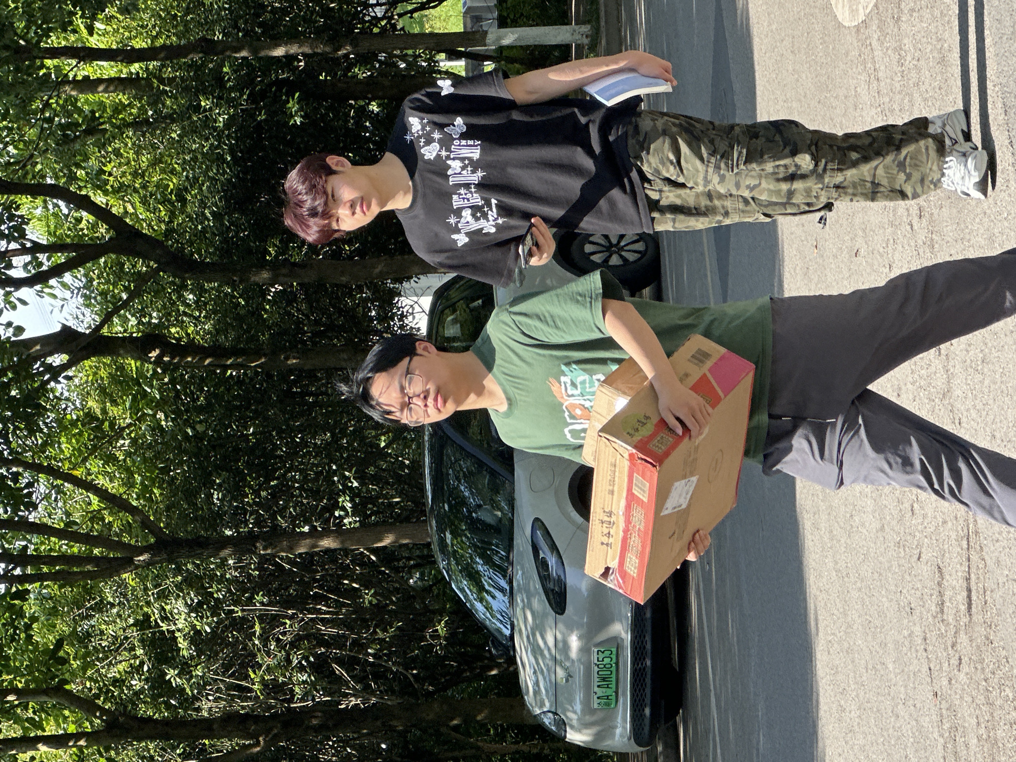 This image captures two young men standing outdoors on a sunny day in Jiangjin, China. The setting features a paved ground, possibly a street or parking area, flanked by dense green trees and foliage, indicating a natural or park-like environment.
In the foreground, a man with dark hair and glasses wears a green t-shirt and dark pants. He is holding a medium-sized cardboard box, which has some red tape or design on it with Chinese characters, possibly "食品" (food) and "特产" (local specialty) on a white label. He looks towards the camera with a neutral expression.
Slightly behind him, to the left, another man with dark red hair is dressed in a black t-shirt featuring white graphic designs including butterflies and abstract shapes, with the brand name "KENZO" visible. He wears camouflage pants and white sneakers. He holds a small, light blue book or notebook in his left hand and a smartphone in his right, also looking towards the camera.
Between the two individuals, a light-colored compact car is partially visible. Its license plate, clearly readable, displays "云 AHD853". The bright sunlight casts distinct shadows on the ground, suggesting it is daytime, either morning or afternoon, with clear weather. Both men exhibit calm and collected demeanors.