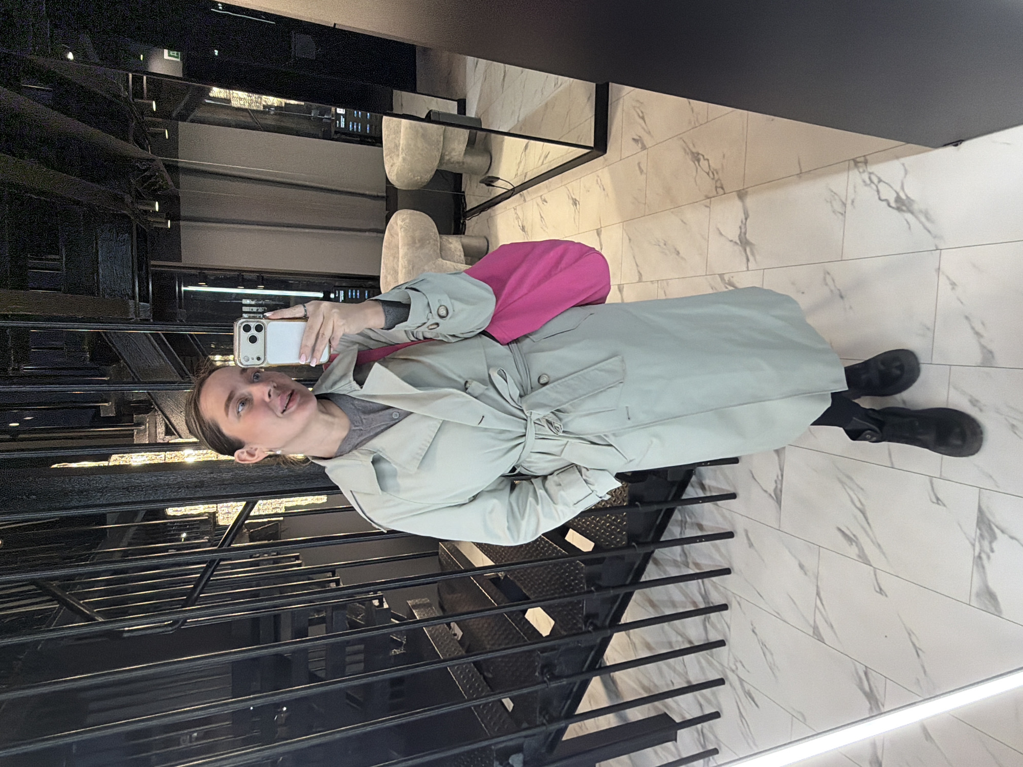 A woman is taking a selfie in what appears to be the Mercury Tower in Moscow, Russia. She is wearing a light green trench coat over a grey top, black leggings, and black boots. She is holding a white cell phone. Behind her is a black metal staircase. The background includes a marble-tiled floor and walls, and seating can be seen in a reflection.