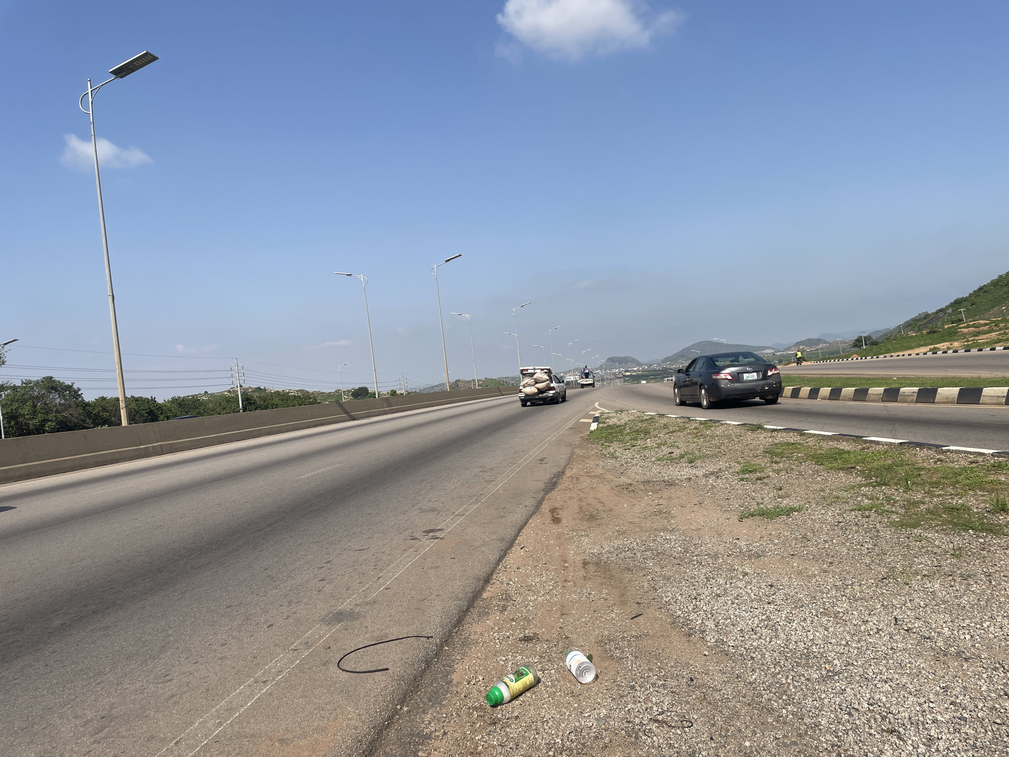 The image depicts a highway scene in Gosa, Nigeria, under a clear blue sky with a few scattered clouds. The time of day appears to be midday, indicated by the bright sunlight casting distinct shadows. The highway stretches into the distance, with multiple lanes visible.
On the left side of the highway, a concrete barrier separates it from an area with trees and utility poles. Several modern streetlights with solar panels are spaced along the roadside. The road surface is asphalt, with white lane markings. On the right side of the highway, there is a gravel shoulder with some sparse vegetation.
Several vehicles are traveling on the highway. In the foreground, a dark-colored car is moving away from the viewer. Further down the road, a white pickup truck is carrying a load of goods on its roof, secured with ropes. A person is riding on the back of this truck. Another car is visible in the distance behind the pickup truck.
On the gravel shoulder in the foreground, there are a couple of discarded items, including a green and white plastic bottle and a white plastic cup, suggesting litter. A black flexible tube or hose is also lying on the asphalt. In the background, rolling hills and more distant settlements can be seen, with lush green vegetation covering the slopes of some hills. The overall impression is one of a busy highway in a semi-urban or developing area. No discernable emotions are conveyed, and the scene is factual and straightforward.