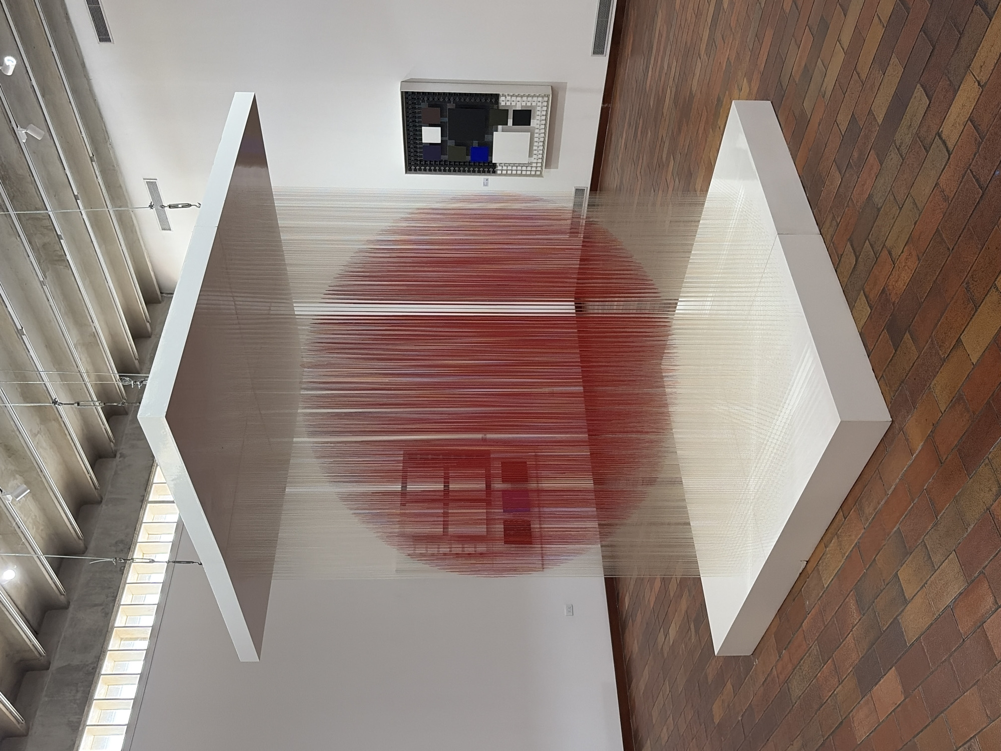 The image shows an indoor art installation. Two white, rectangular prism structures with pointed ends face each other, seemingly suspended in air. Between them, a dense arrangement of string creates a circular, reddish-brown form. The strings appear to be taut between the two prisms. Behind the installation, a white wall displays a square artwork composed of smaller squares with various colors and patterns. The architectural setting includes a concrete ceiling with linear elements and windows, and a brick wall. The location is most likely an art museum or gallery. The information about the city of Ciudad Bolivar, Venezuela is not evident from the image itself.