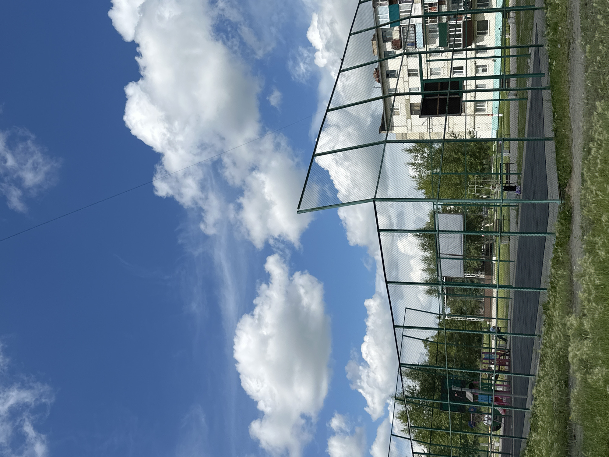 The image depicts a fenced-in outdoor sports court, possibly a basketball court, with a playground visible within its confines. The court has a dark, textured surface. Beyond the court, trees form a backdrop, and a multi-story apartment building is situated further back. The sky is blue with scattered white clouds, and a thin wire runs diagonally across the upper portion of the frame. In the foreground, tall, feathery grass grows. There are several people present: two figures are on the playground equipment, appearing to be children, and one person is sitting on a bench on the court. The scene is likely located in Rudny, Kazakhstan, as indicated by the architectural style of the apartment building and the general setting.