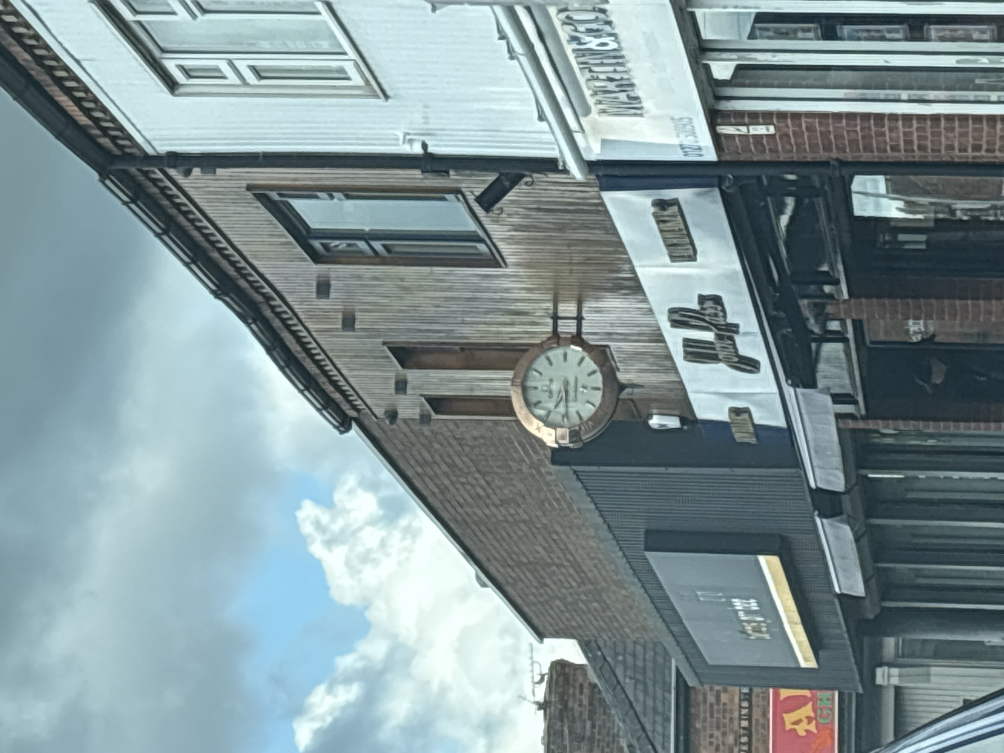 This image shows the exterior of the bar/restaurant "Tom's Tap" located in Crewe, United Kingdom. An outdoor clock hangs on the building's side, next to the sign. Above, a mix of brick and wooden paneling is visible. The sky is overcast with patchy clouds.