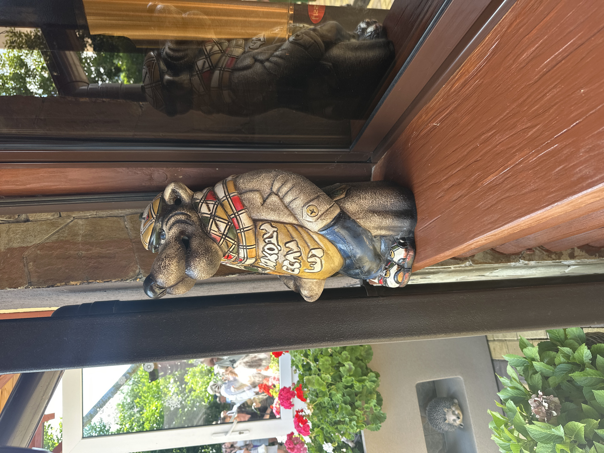 A statue of a badger in traditional clothing stands on a wooden windowsill in front of a window reflecting trees and people. The statue has a patterned scarf and a yellow tunic with writing on it. In the background, through the window, a group of people are visible, as well as potted flowers. A small hedgehog figurine is also visible on a shelf below the windowsill. The scene appears to be outdoors, possibly at a cafe or shop in Krasnaya Polyana, Russia, given the architectural style and vegetation.