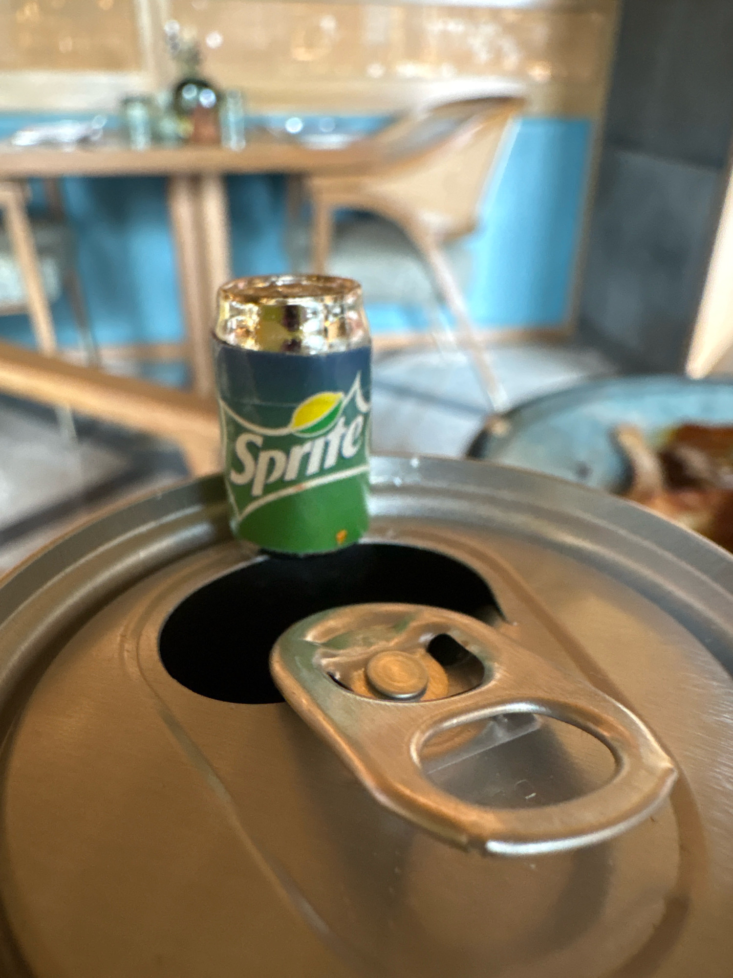 A macro shot of an opened aluminum can lid, with a miniature Sprite can perched on top. The can lid is a dull silver color, with a raised circular opening and a silver tab. The Sprite can is green and white, with the Sprite logo clearly visible. The background is blurred and shows a restaurant setting, with tables, chairs, and a blue wall. The image appears to be taken indoors, likely in a restaurant in Vagator, India. There are no people or animals visible in the image. The overall impression is a playful and whimsical scene, with the miniature can adding a touch of novelty.