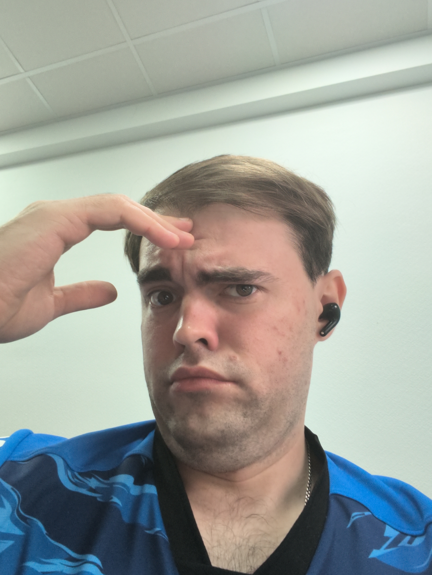A man is pictured in a close-up, head and shoulders shot. He has light brown hair and fair skin with some blemishes. His left hand is raised to his forehead, obscuring part of his brow. He is wearing black earbuds in his right ear and a blue shirt with a black collar. The background is a plain, light-colored wall with a tiled ceiling visible at the top. The lighting appears to be overhead.