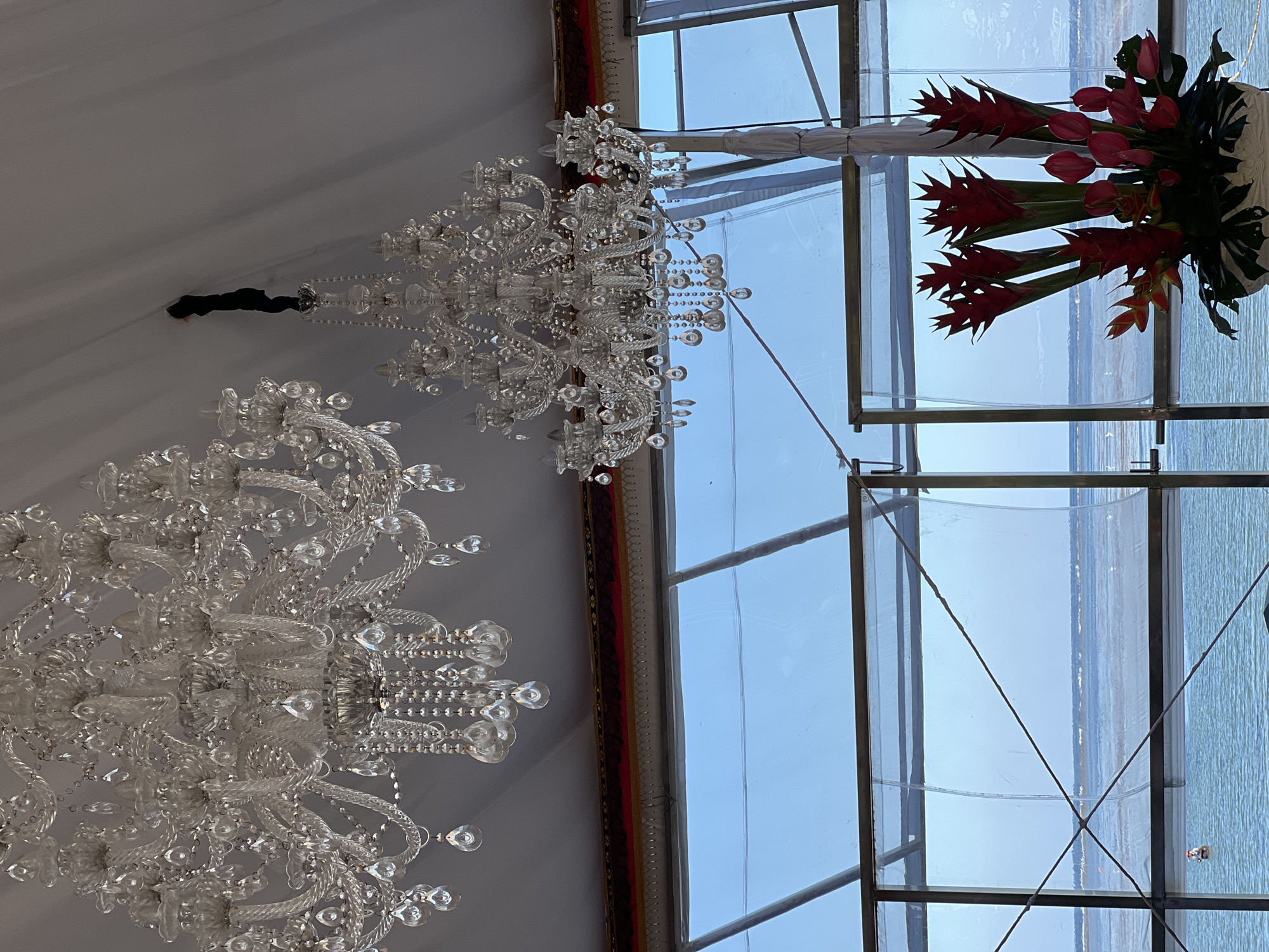 The image shows the interior of a tent-like structure in Denpasar, Indonesia. Two elaborate crystal chandeliers hang from the ceiling. A large floral arrangement sits atop a metal support structure with translucent panes behind it, offering a view of a body of water outside. The sky is visible through the panes.