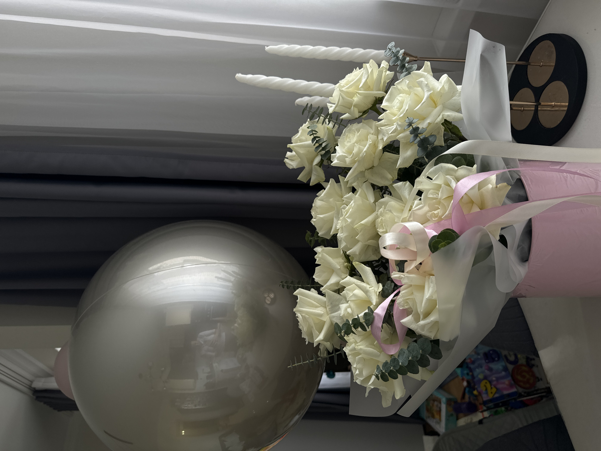 The image displays an indoor scene set for a celebration, featuring a bouquet of white roses and a large silver-grey balloon. The bouquet, centrally placed on the right side, consists of numerous pristine white roses accented with sprigs of green eucalyptus. It is beautifully wrapped in a combination of clear film and light pink paper, tied with elegant light pink and white ribbons.
To the left, a large, spherical metallic silver-grey balloon is prominently featured, its reflective surface mirroring the surrounding room, including indistinct indoor structures. In the upper background, three tall, white twisted candles are visible, suggesting a celebratory cake or display. Adjacent to the candles, on a wall or stand, is a circular black decorative piece adorned with three golden circular elements and slender golden rods. The left background is filled with layered grey curtains or drapes, transitioning from lighter to darker shades. Below the bouquet, a colorful item resembling a decorated phone case or a small board game is partially visible.
The arrangement of flowers, a balloon, and candles strongly implies a special occasion, such as a birthday or anniversary. The setting appears to be brightly lit, possibly by natural light during the daytime. No legible text is present in the image. The location context provided is Akulovo (Gp Kubinka), Russia.