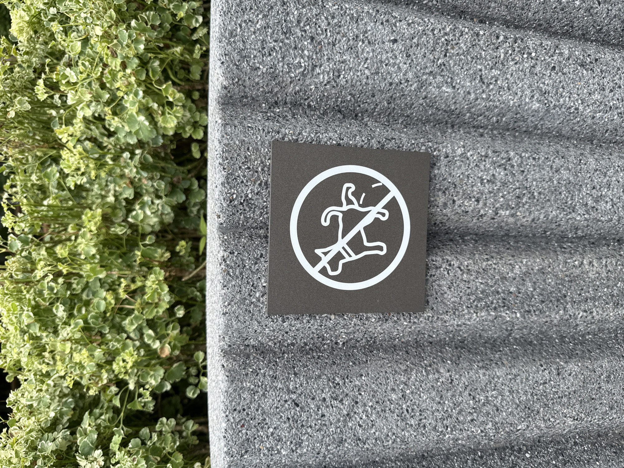 The image shows a sign with a white circle and a diagonal line over a silhouette of a dog relieving itself. The sign is affixed to a textured gray concrete bin. Behind the bin is lush green foliage. The bin appears to be located outdoors, possibly in a park or public space. The sign indicates that dogs are not allowed to urinate or defecate in this area. The overall scene is calm and orderly, with no people or animals present. The image is likely taken in Bangkok, Thailand, based on the presence of the specific type of foliage and the commonality of such signage in public areas.