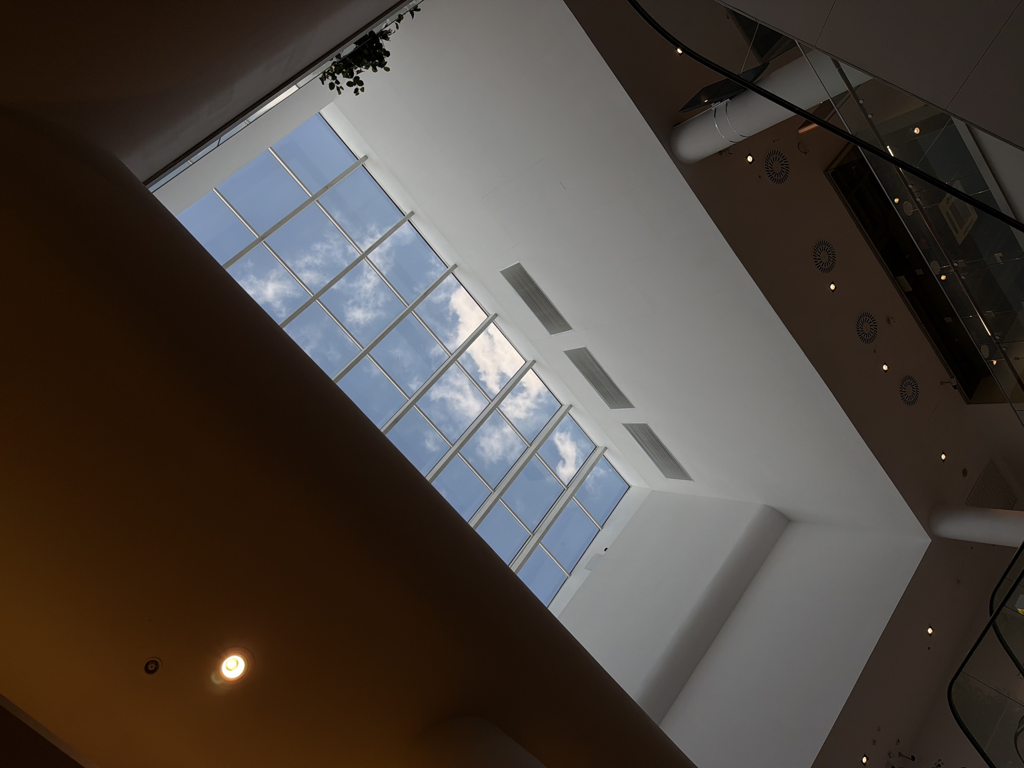 The image captures an upward-looking perspective within a modern building, likely a shopping mall or a large public atrium, characterized by its contemporary architecture and dramatic lighting. A prominent feature is a large, gridded skylight in the upper left section of the frame, revealing a clear blue sky with scattered white clouds, indicating it is daytime.
The ceiling structure is multi-faceted and angled. A significant portion in the foreground and lower-left is a warm, dark brown/orange color, featuring at least one illuminated recessed light fixture. This darker ceiling contrasts sharply with the bright white and light grey sections of the ceiling that extend towards the upper and right sides of the image. These lighter ceiling areas are equipped with rectangular air vents and several circular ventilation grilles, along with additional recessed lighting fixtures.
In the upper-left corner, a small cluster of dark green hanging foliage is visible, adding a touch of nature to the otherwise sleek interior. On the far right, reflections on what appear to be glass panels and black handrails suggest the presence of an escalator or a multi-level structure. No people are visible, and no specific activity is taking place, focusing instead on the architectural space itself. The overall impression is one of spaciousness and modern design, illuminated by both natural daylight and artificial light sources. No text is visible in the image.