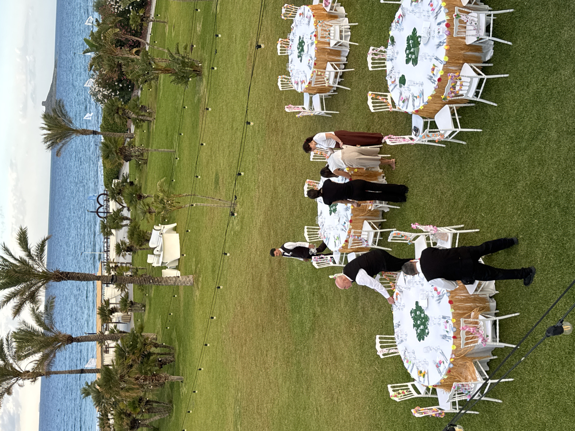 This aerial view captures an outdoor event setup on a lush green lawn adjacent to the sea. Several round tables, draped in white tablecloths adorned with festive, colorful leis and foliage, are arranged on the grass. White chairs, also decorated with leis, surround the tables. In the background, palm trees and other vegetation frame the scene, with the tranquil blue sea extending to the horizon. A distant structure, possibly a historical tower, is visible on a rocky outcrop in the sea.
Several individuals, dressed in formal attire including vests and shirts, appear to be attending to the tables, suggesting preparation for a gathering or event. The lighting string with bulbs suggests the event may extend into the evening. The location is Isola delle Femmine, Italy, a coastal town renowned for its natural beauty and historic landmarks.