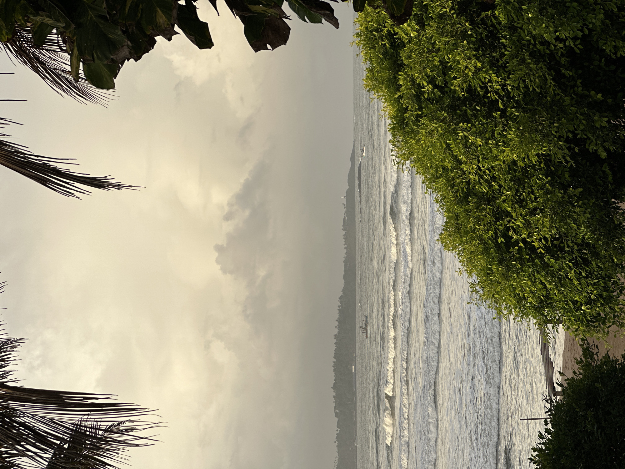 This image depicts a coastal scene in Peleana, Sri Lanka, likely during late afternoon or early morning due to the diffused light. The foreground is dominated by lush green foliage, with palm fronds framing the top of the image. In the middle ground, a body of water with gentle waves stretches towards a distant tree-lined coastline. Two small boats are visible on the water, one closer to the shore and another further out. A lighthouse can be seen on a promontory in the far distance. The sky is overcast with scattered clouds, lending a soft, atmospheric quality to the scene. The beach area in the lower part of the image shows some sand and a wooden structure, possibly a pier or walkway. No people are clearly visible.