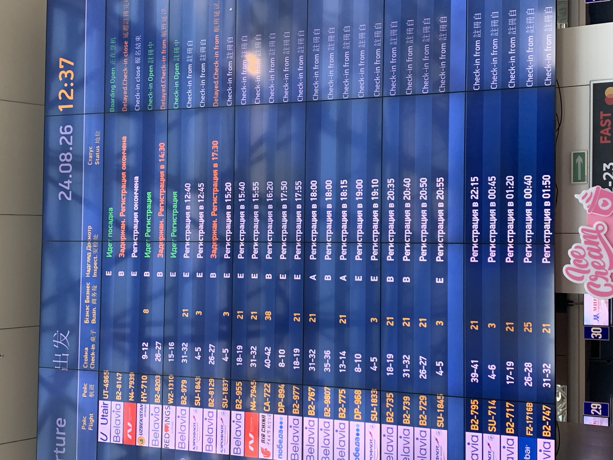 The image is a photograph of a departure board at Minsk National Airport (MSQ), Belarus, on August 24, 2024, at 12:37. The board displays flight information in Russian and English, including flight numbers, destinations, check-in counter numbers, business class check-in, and flight status. Several flights are listed with various statuses such as "Boarding Open," "Delayed," and "Registration." The environment appears to be an indoor airport terminal, with the lighting typical of such a setting. There are no people clearly visible in the frame, but the context suggests a busy airport environment. In the foreground, partially visible, are elements of advertising, including an "Ice Cream" sign and some text that includes "-23 FAST."