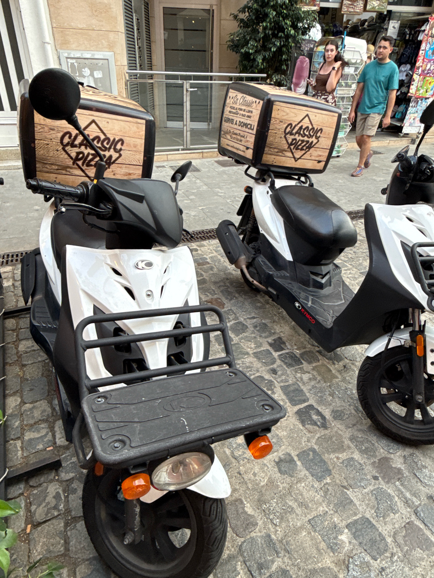 Two white Kymco scooters, each equipped with a wooden-style pizza delivery box, are parked on a cobblestone street in Blanes, Spain. The scooter in the foreground is positioned at a slight angle, facing towards the right of the frame, with its black seat and white front panel clearly visible. The rear of this scooter features a black rack with a license plate holder and an orange turn signal. The scooter in the background, to the right, is mostly obscured by the scooter in the foreground, but its black seat and white front panel are discernible.
To the left of the scooters, a building with light-colored walls and shuttered windows is visible. In the background, a man and a woman are walking on the street. The man, wearing a teal t-shirt and khaki shorts, walks towards the left, while the woman, in a brown top and floral skirt, is looking towards the left. Various shop displays are visible behind them, suggesting a commercial area. The ground is paved with textured cobblestones, and some greenery is present on the left side of the image.
