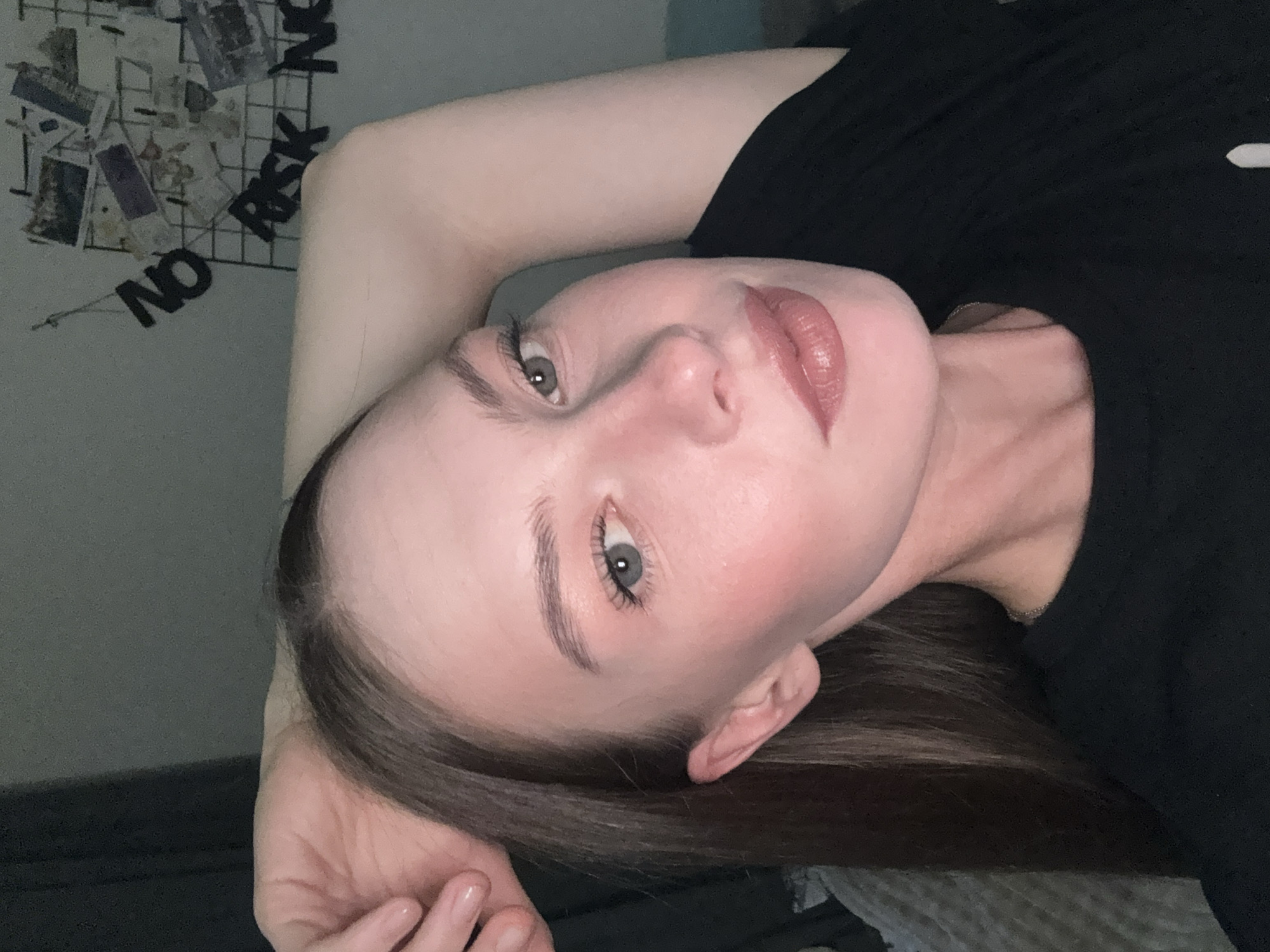 This is a close-up, low-angle, portrait-style photograph of a young person, likely female, with fair skin and dark brown hair. The person is lying down, resting their head on their left arm, and looking directly at the camera. Their head is tilted, and the image is oriented such that the person's face appears to be at the top of the frame, though the full image suggests the person is lying on their side.
The person is wearing a black t-shirt. Their makeup includes foundation, blush on the cheeks, mascara, and a lip gloss in a muted pink shade. Their eyebrows are well-defined. The background is out of focus, but it appears to be an indoor setting. To the upper left of the frame, there is a wire grid with several small photos and some black letter cutouts that spell out "NO RISK". The lighting is soft, suggesting it might be evening or a dimly lit room. The overall mood conveyed is casual and relaxed, possibly intimate due to the close framing and direct gaze. The location context of Perm, Russia, does not provide any specific visual cues within the image itself.