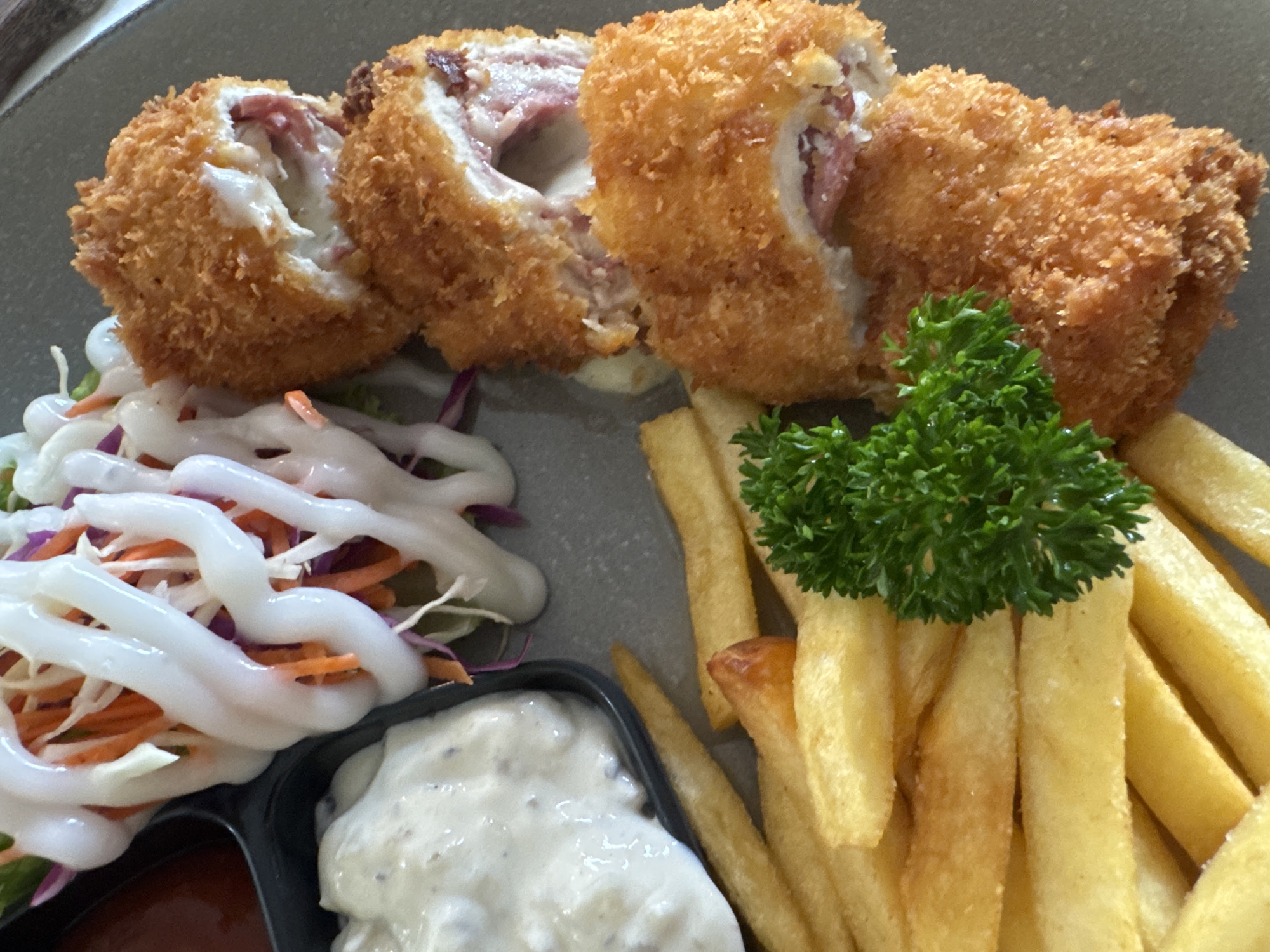 A close-up shot depicts a plate of food. The main component is sliced, breaded, and fried chicken cordon bleu. The chicken appears to be stuffed with ham and cheese. The dish is accompanied by French fries, a small salad with white dressing, and two dipping sauces in a divided container. The scene appears to be set in Kuta, Indonesia.