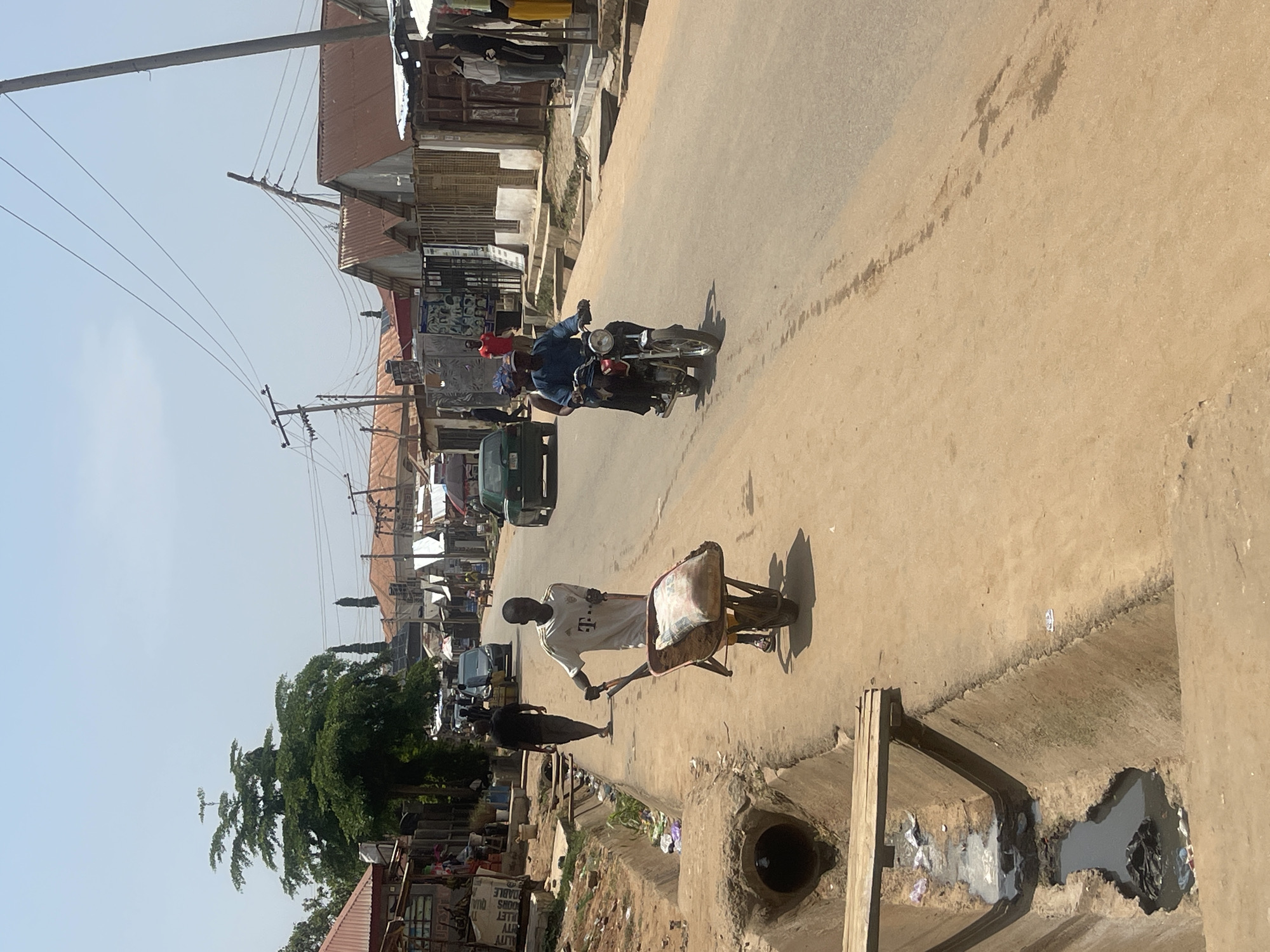 The image depicts a street scene in Abuja, Nigeria, likely a commercial or residential area, under a bright, hazy sky. A young man wearing a white t-shirt with a prominent "T" logo is pushing a wheelbarrow filled with a large bag of cement or similar material along a dusty dirt road. To his left, a concrete drainage ditch runs alongside the road, containing murky water and debris, with a makeshift wooden plank serving as a small bridge over a section of it. In the background, a few vehicles are present, including a green car and a motorcycle carrying two people. The buildings lining the street are varied, with some appearing to be shops with corrugated metal roofs and others with more permanent structures. Electrical poles and overhead wires crisscross the sky. The overall impression is of a bustling, ordinary day in a Nigerian city, with elements of commerce, transportation, and infrastructure typical of such an environment.