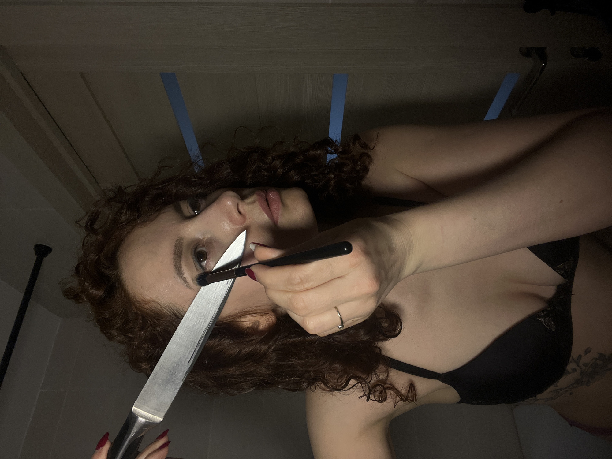 A woman with curly red hair, wearing a black bra, is holding a kitchen knife and a makeup brush near her eye. She has red nail polish and a ring on her finger. The background appears to be a bathroom with a white door and blue tape. The specific location within the city of Saint Petersburg, Russia, cannot be determined from the image.