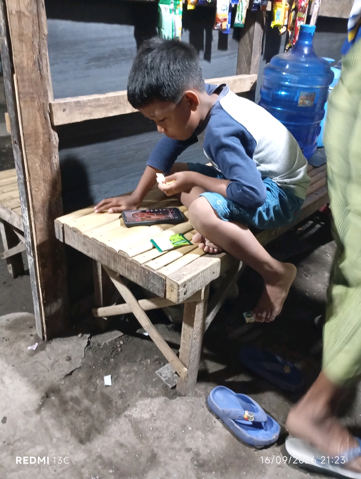 A young boy with dark hair is seated on a wooden bench, engrossed in looking at a smartphone. He is wearing a blue and white long-sleeved shirt and blue shorts. His left hand is on the bench, near the phone, while his right hand holds a small white object, possibly food or candy. A pack of green candy is also on the bench. To the right of the boy, a large blue water jug sits on a shelf behind the bench, and an assortment of packaged goods are displayed above. The bench is situated in an outdoor area with a concrete floor. A pair of blue flip-flops are on the ground near the bench, and another person's legs and feet, wearing green pants and white flip-flops, are visible on the right side of the frame. The scene suggests a small, informal shop or stall. The timestamp at the bottom right indicates the photo was taken on 16/09/2026 at 21:23. The location is identified as Wuluhan, Indonesia.