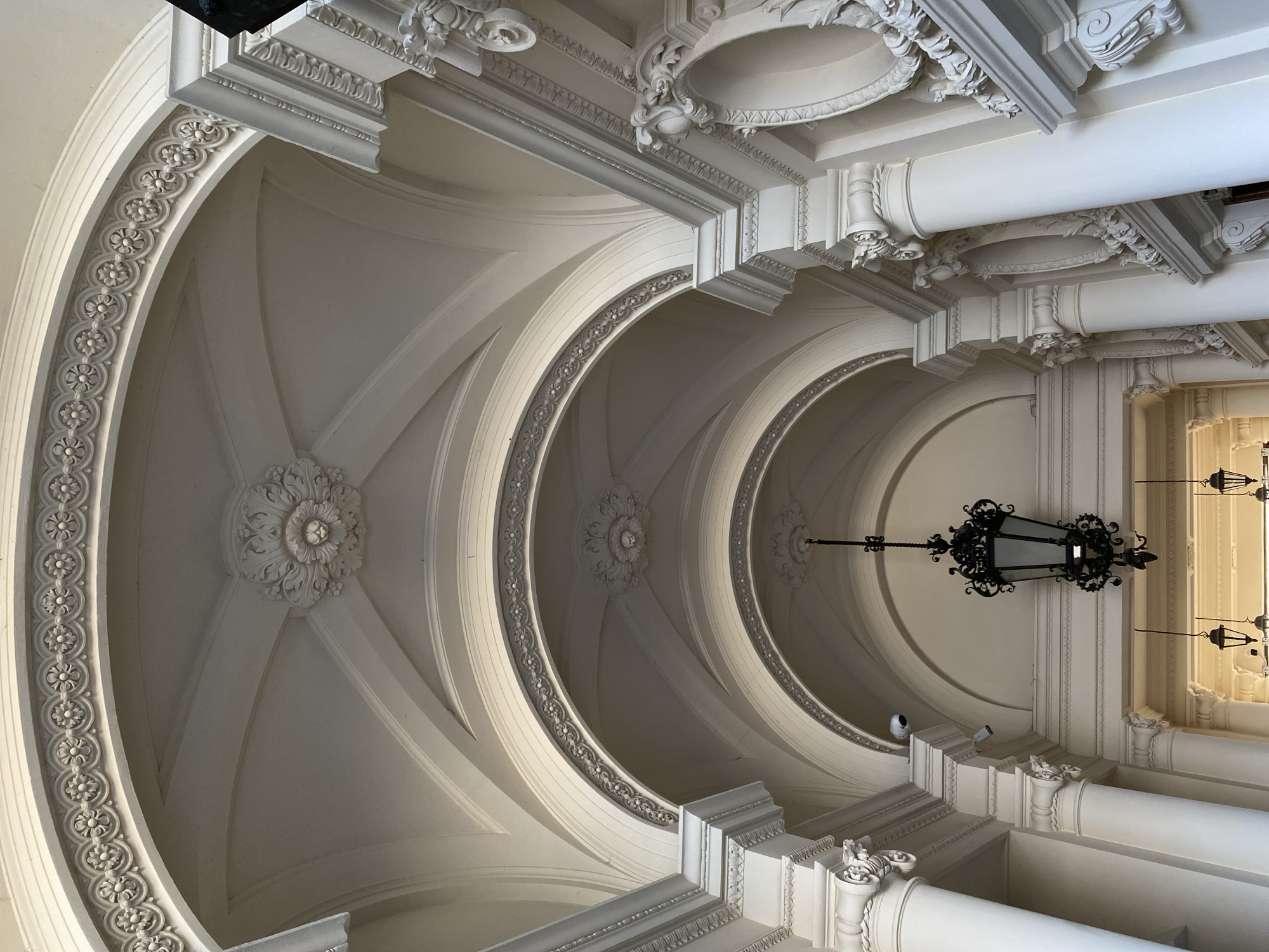 This is a low-angle, close-up shot of the ornate ceiling and upper walls of an interior architectural space in Warsaw, Poland. The architecture features multiple arched passageways with intricate decorative molding, including repeating floral patterns along the edges of the arches and elaborate rosettes at the apex of some ceiling sections. White classical columns with detailed capitals support the structure. A large, black, ornate lantern hangs from the ceiling in the middle of the frame. Smaller, similar lanterns are visible further down the passageway. Two security cameras are mounted on the upper wall to the left. The lighting is soft, creating subtle shadows that highlight the architectural details. The scene is devoid of people.
