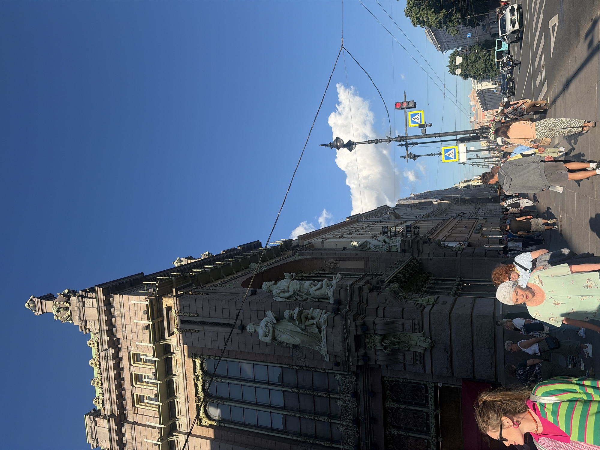 The image depicts a street scene in Saint Petersburg, Russia, likely on a sunny day given the clear blue sky and the shadows. In the foreground, a woman with blonde hair tied in a ponytail, wearing sunglasses and a brightly colored striped shirt, is looking to her left. Adjacent to her, an older woman in a light green patterned blouse and white head covering is walking towards the viewer.
The background is dominated by a large, ornate building with classical architectural elements, including statues on its facade and detailed stonework. The building appears to be a significant structure, possibly a historical landmark or a public building. Across the street, a busy road is visible with several cars, a bus, and a motorcycle. Traffic lights and pedestrian crossing signs are present, indicating an intersection. People are walking on the sidewalk and crossing the street, suggesting a lively urban environment. The architecture and atmosphere are characteristic of Saint Petersburg's grand boulevards.
