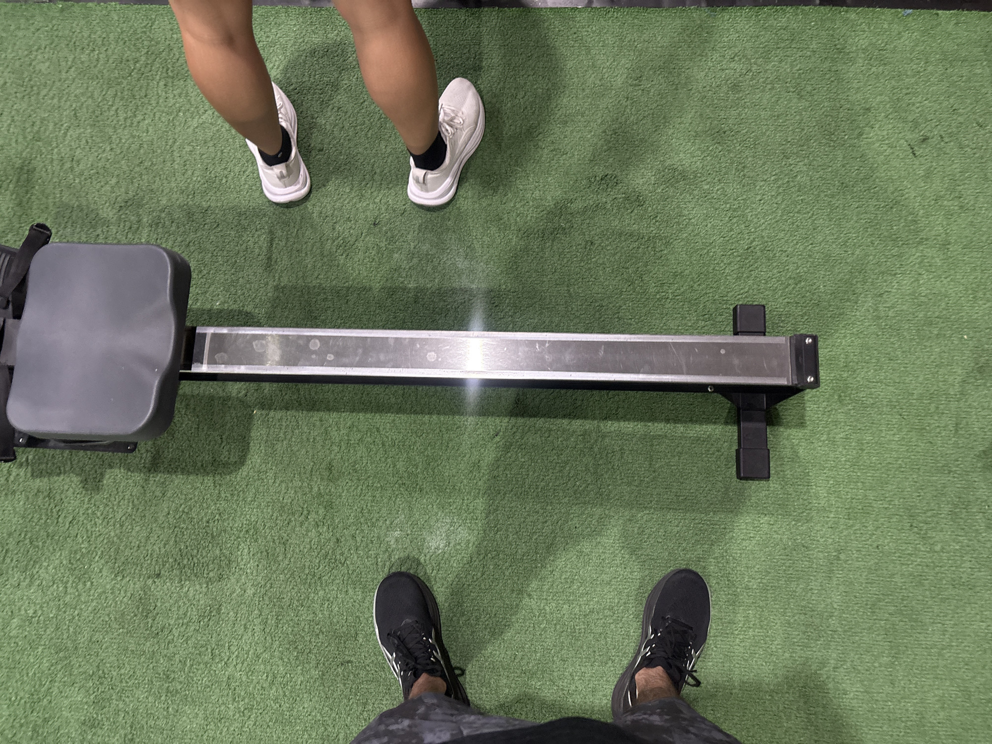 The image shows an overhead view of a rowing machine on a green turf surface. Two people are visible, one at each end of the machine. The person at the front of the machine is wearing white sneakers and black socks. The person at the back is wearing black sneakers and patterned shorts. The rowing machine is silver and black. The location is presumed to be a gym or fitness area. The location is unknown.