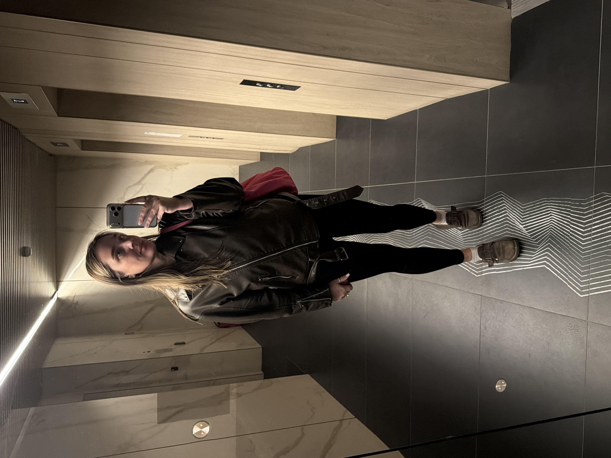 A woman stands in an elevator, taking a selfie with a smartphone. She wears a brown leather jacket, black leggings, and brown boots. The elevator interior features marble-like walls and light wood paneling. The floor is dark with a decorative geometric pattern. The location is Moscow, Russia.