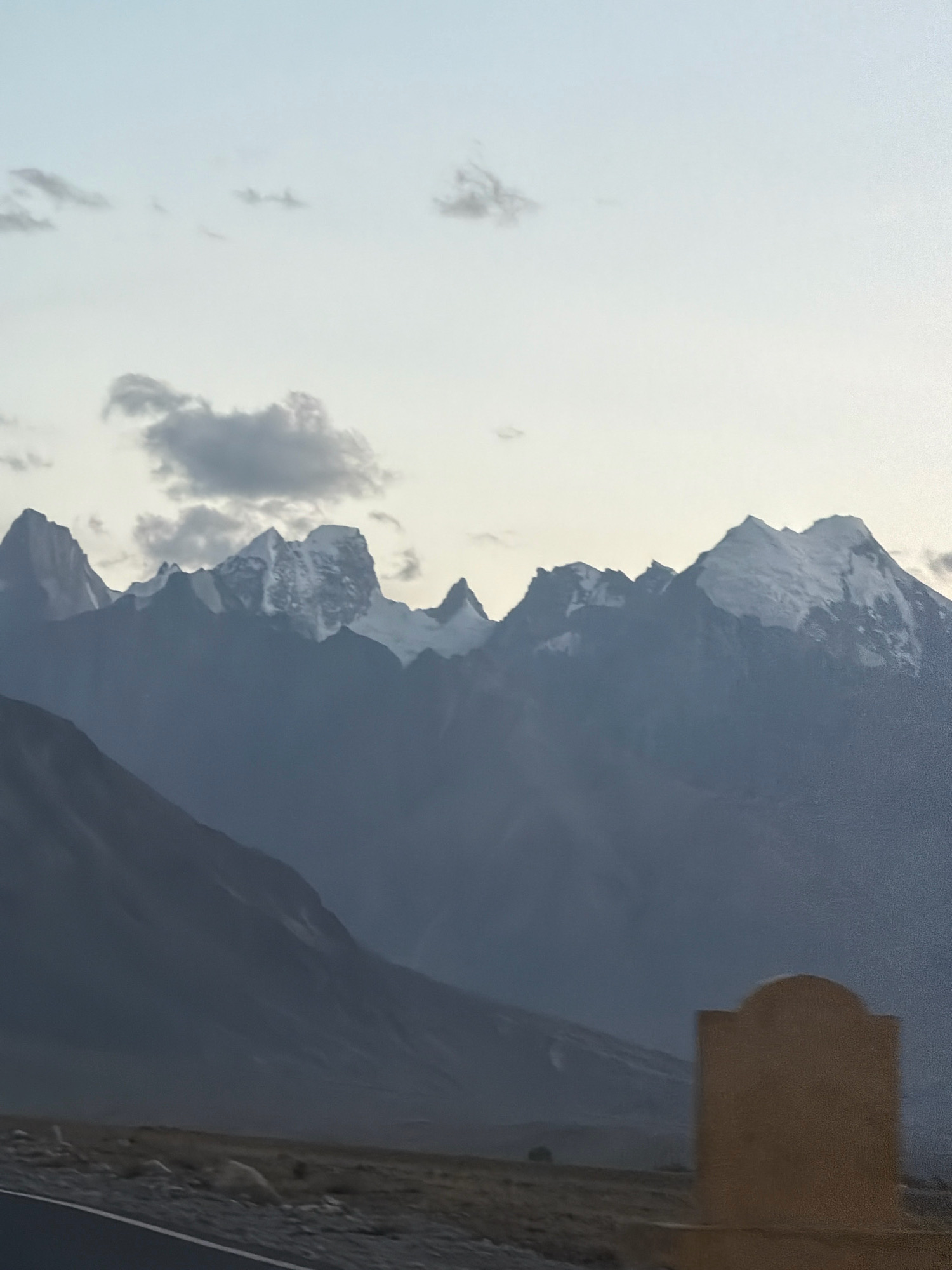 A wide shot captures a mountain range in the distance under a hazy sky. The mountains have jagged, snow-capped peaks. In the foreground, a dark, sloping mountain or hill is visible, with a sparsely vegetated, rocky landscape below. A paved road runs along the bottom left of the image. A blurred, tan-colored structure with a curved top is in the right foreground. The scene appears to be in Stongde, India, given the geographical context of the image.