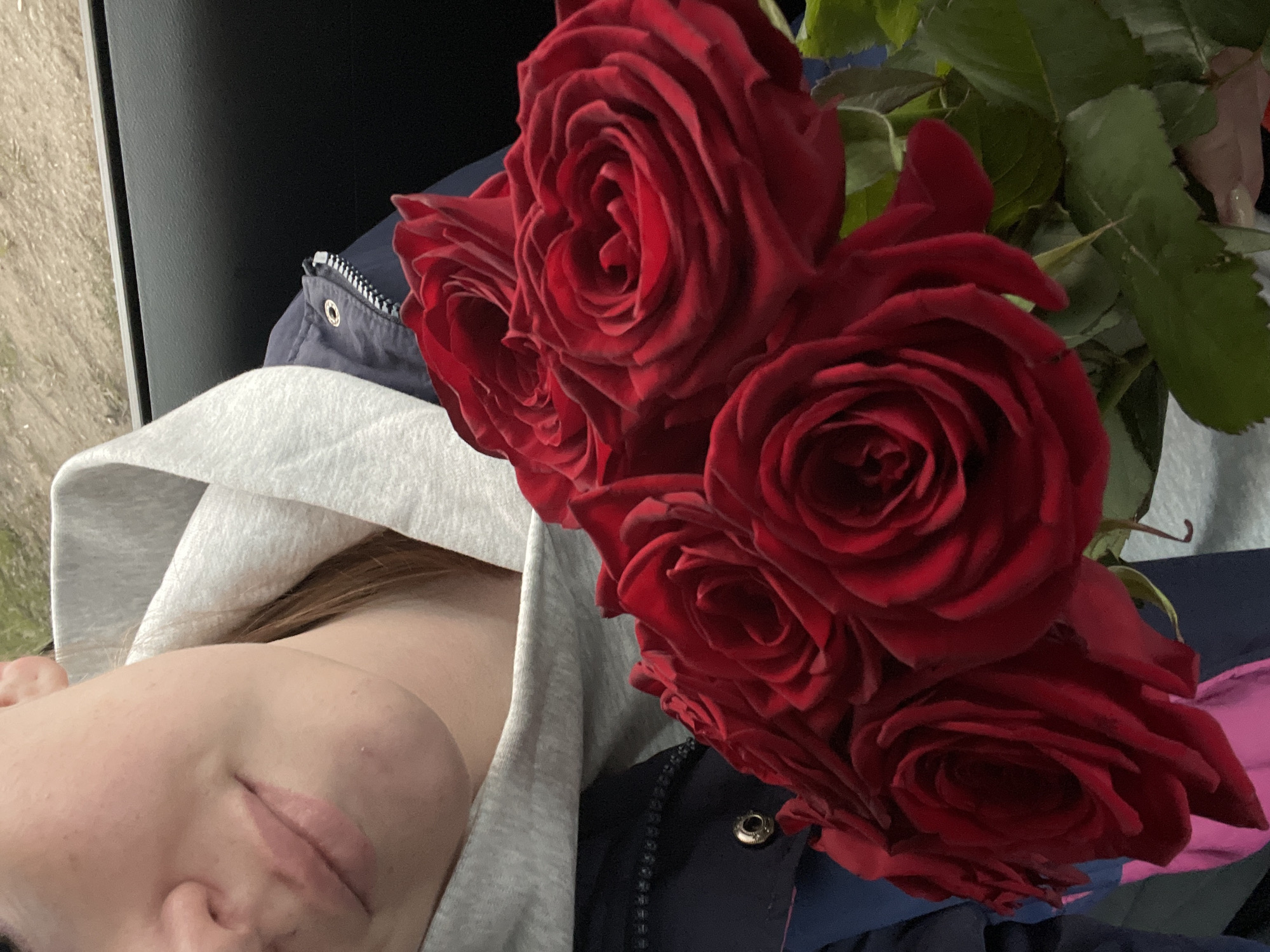 The image shows a close-up, angled view of a person resting alongside a bouquet of red roses, potentially in Chaikovsky, Russia. The person's face is partially visible on the left side, showing their chin, lips, nose, and part of their cheek and forehead. They appear to be lying down, with a serene and relaxed expression, possibly with their eyes closed or looking away from the camera. They are wearing a light grey hooded sweatshirt.
Dominating the right side of the frame is a vibrant bouquet of at least six large, deep red roses with prominent green leaves. The roses are in full bloom, showcasing their velvety petals. Beneath the roses and next to the person, a dark blue garment, possibly a jacket or coat, is visible, featuring a silver zipper and a circular metal eyelet.
The background behind the person and flowers is largely dark and indistinct, suggesting an interior setting such as a car or a dimly lit room. On the far left edge, a textured, light-colored surface, possibly gravel or pavement, is faintly visible and blurred, implying an outdoor element or a view through a window. There is no visible text in the image.