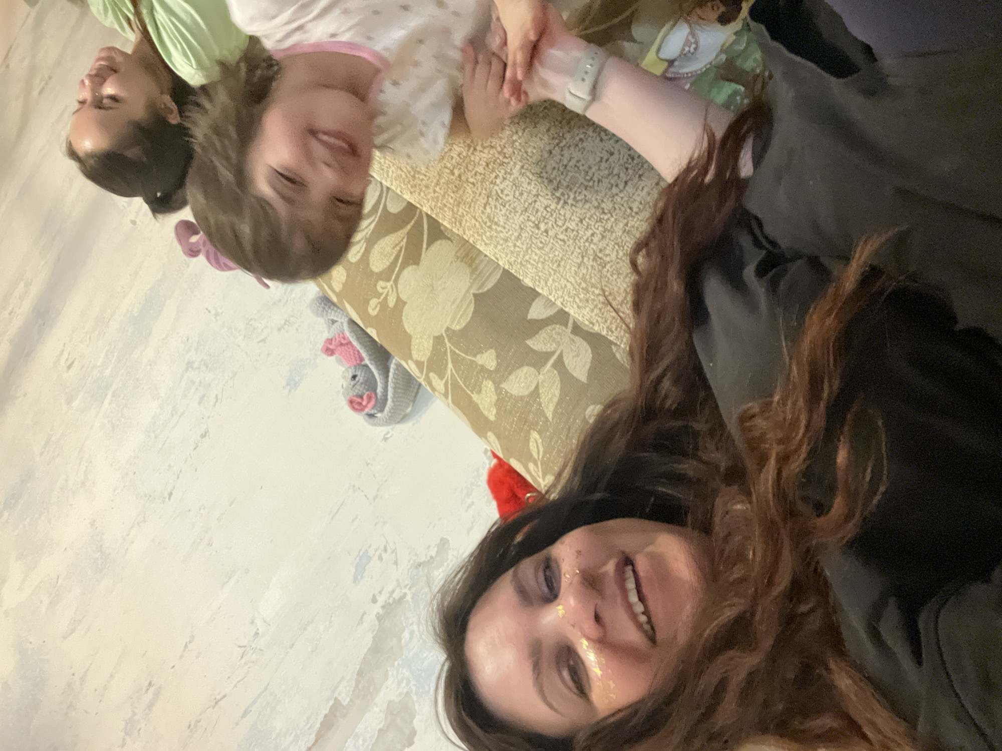 The image shows three individuals—two women and a child—lying together on what appears to be a floral-patterned sofa or bed. The camera is likely held by the woman with long, brown, wavy hair who is visible in the foreground with a full smile. She has light skin, brown eyes, and some gold glitter on her face.
Next to her, on the left side of the image, there is another woman with dark hair pulled back into a bun, wearing a light green shirt.
The child, who appears to be a young girl with short brown hair, is wearing a light-colored pajama set with a pink trim. The child and one of the women seem to be holding hands. The location is Noviy, Russia.
The background includes the corner of a light-colored wall, a gray knitted toy, and a portion of a watch on one of the adult’s wrists. A black garment is visible in the background.