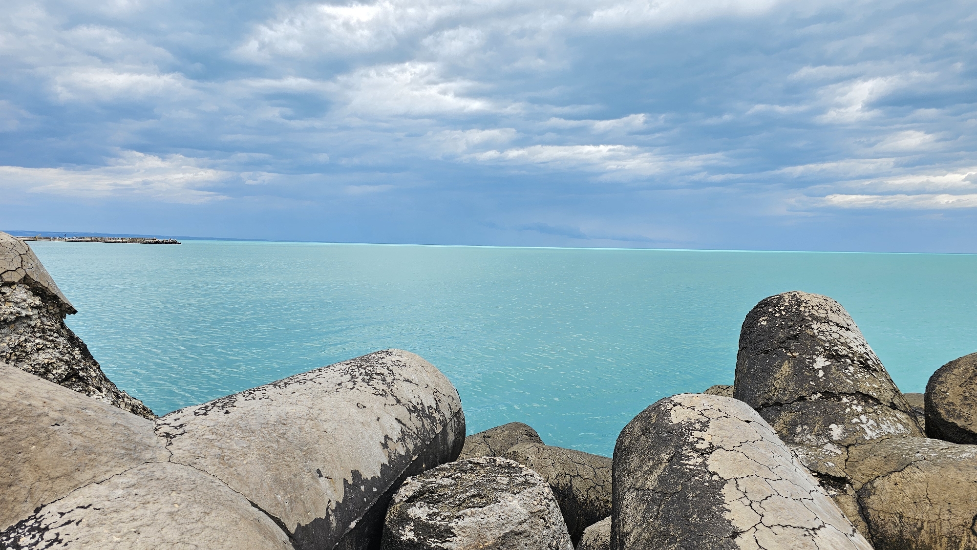The image depicts a coastal scene in Varna, Bulgaria, featuring a breakwater constructed of cylindrical concrete blocks. The turquoise-colored sea extends to the horizon under a cloudy sky. A distant structure, possibly a pier or another part of the port infrastructure, is visible in the far left of the frame. The concrete blocks in the foreground are weathered and show signs of age and exposure to the elements, with cracks and discoloration evident on their surfaces. The focus is primarily on the breakwater and the sea, with the sky filling the upper part of the image. No people are visible in the scene.