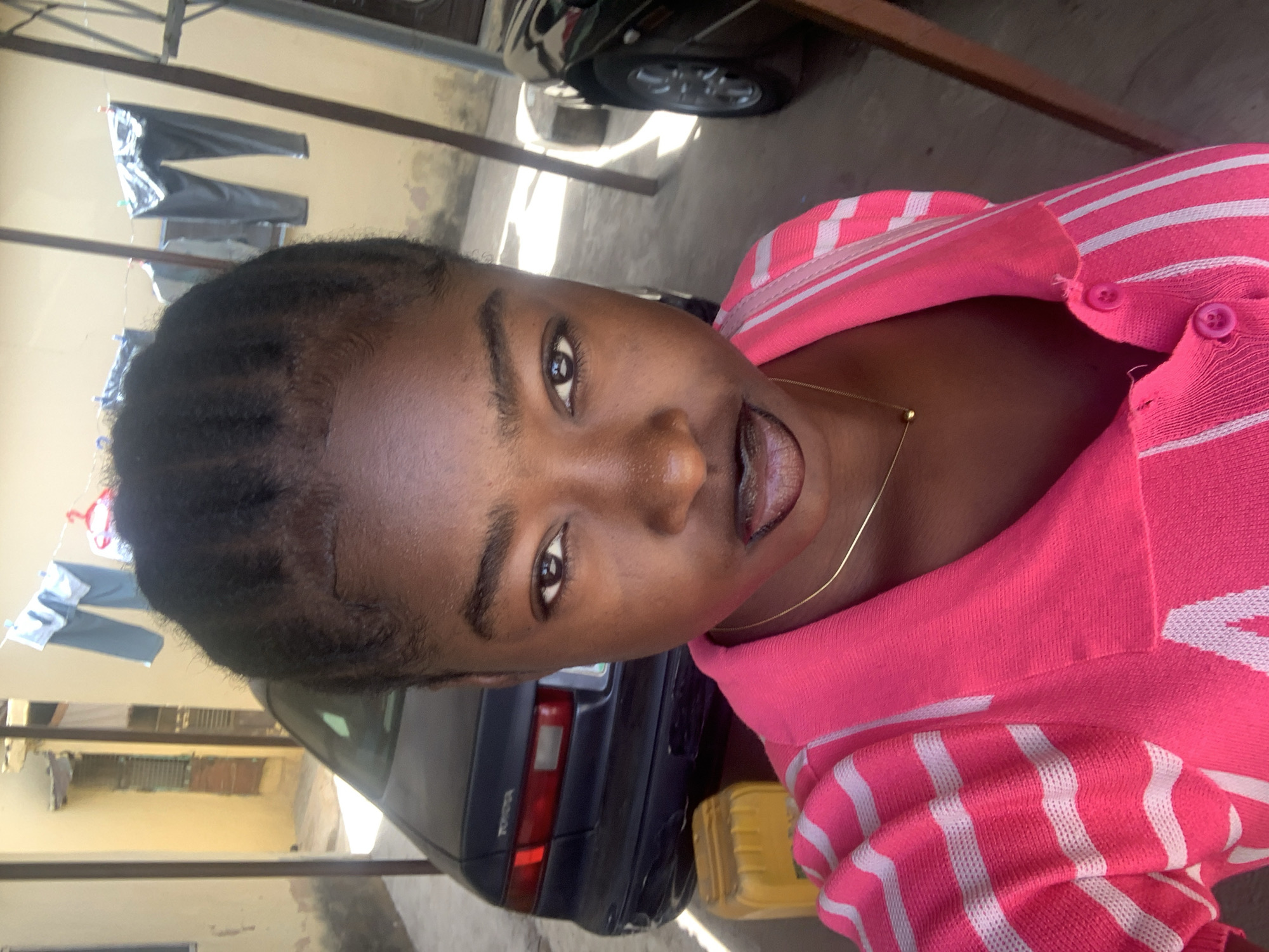 A young, dark-skinned woman with a short, dark hairstyle is centered in the image, wearing a pink and white striped collared shirt. She has a gold necklace around her neck. Behind her is a dark blue sedan, parked on a concrete surface with patches of dirt. In the background, clothes hang on a clothesline attached to a tan-colored building. The location is Rafin Par, Nigeria.