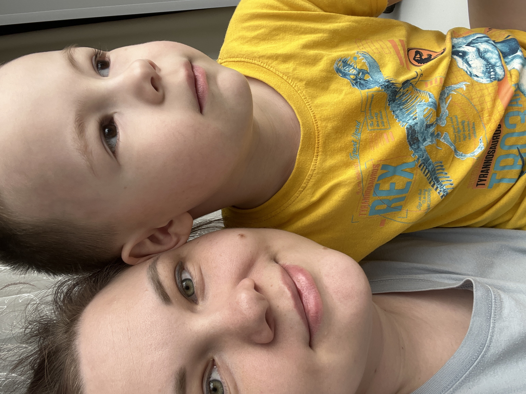A selfie-style, close-up photograph shows a fair-skinned woman and a young boy lying down. The woman is in the foreground, wearing a light grey t-shirt. The boy is positioned above her, wearing a yellow t-shirt with a dinosaur design. Their faces are near each other. The photo was seemingly taken indoors. The location in the city of Usman, Russia, is not deducible from the provided image.