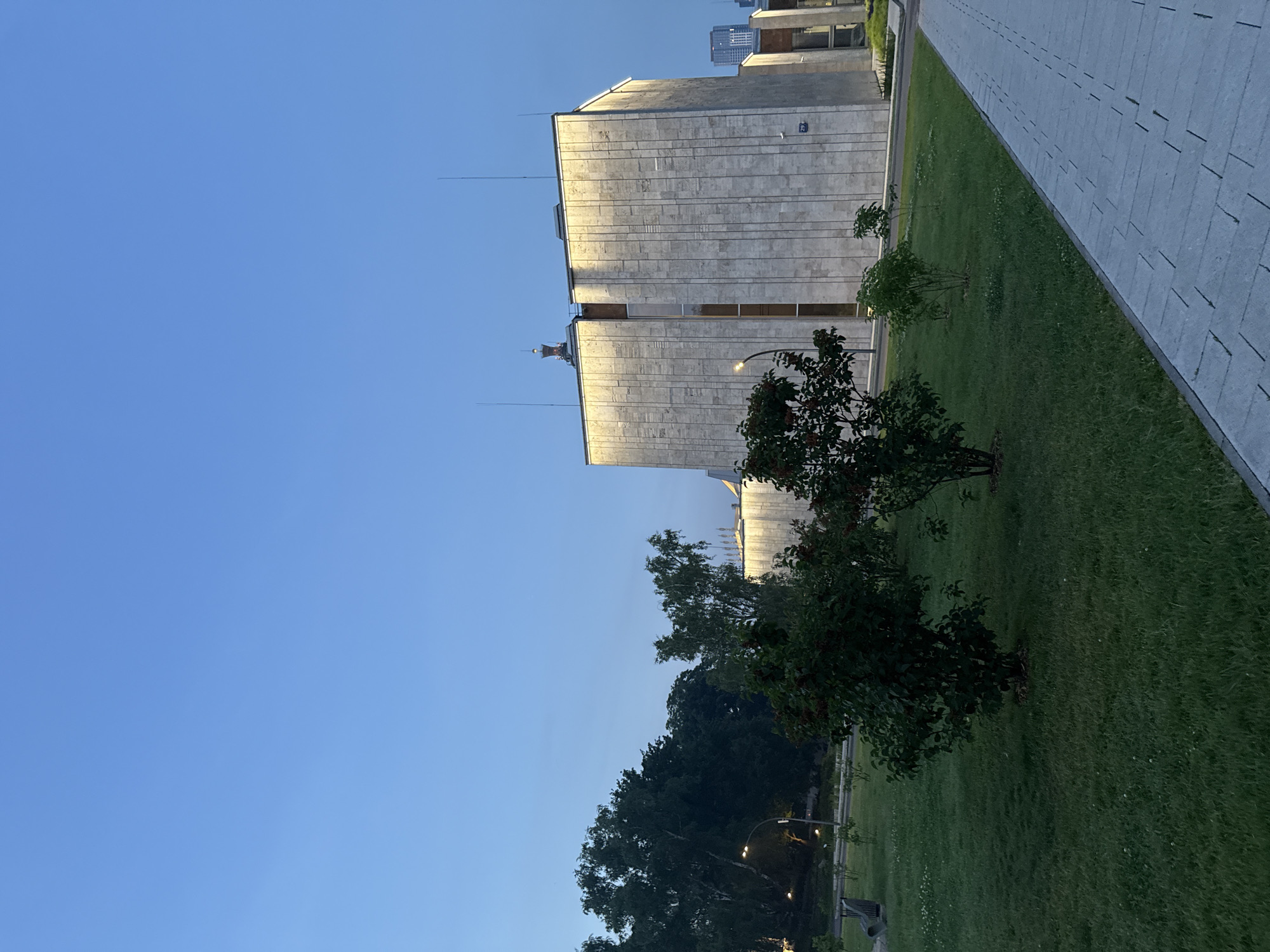The image depicts the building of the Russian Academy of Sciences (RAS) in Moscow, Russia, at dusk. The building is a large, modern structure with a facade clad in light-colored stone panels. The upper section of the building is illuminated by warm, artificial lighting. In the foreground, a well-maintained lawn with several small trees and bushes is visible. A paved walkway runs along the right side of the frame. The sky is a clear, deep blue, indicating twilight. Trees and greenery line the left side of the image, with a streetlamp visible among them. The overall scene suggests a calm and serene evening in an urban park setting.