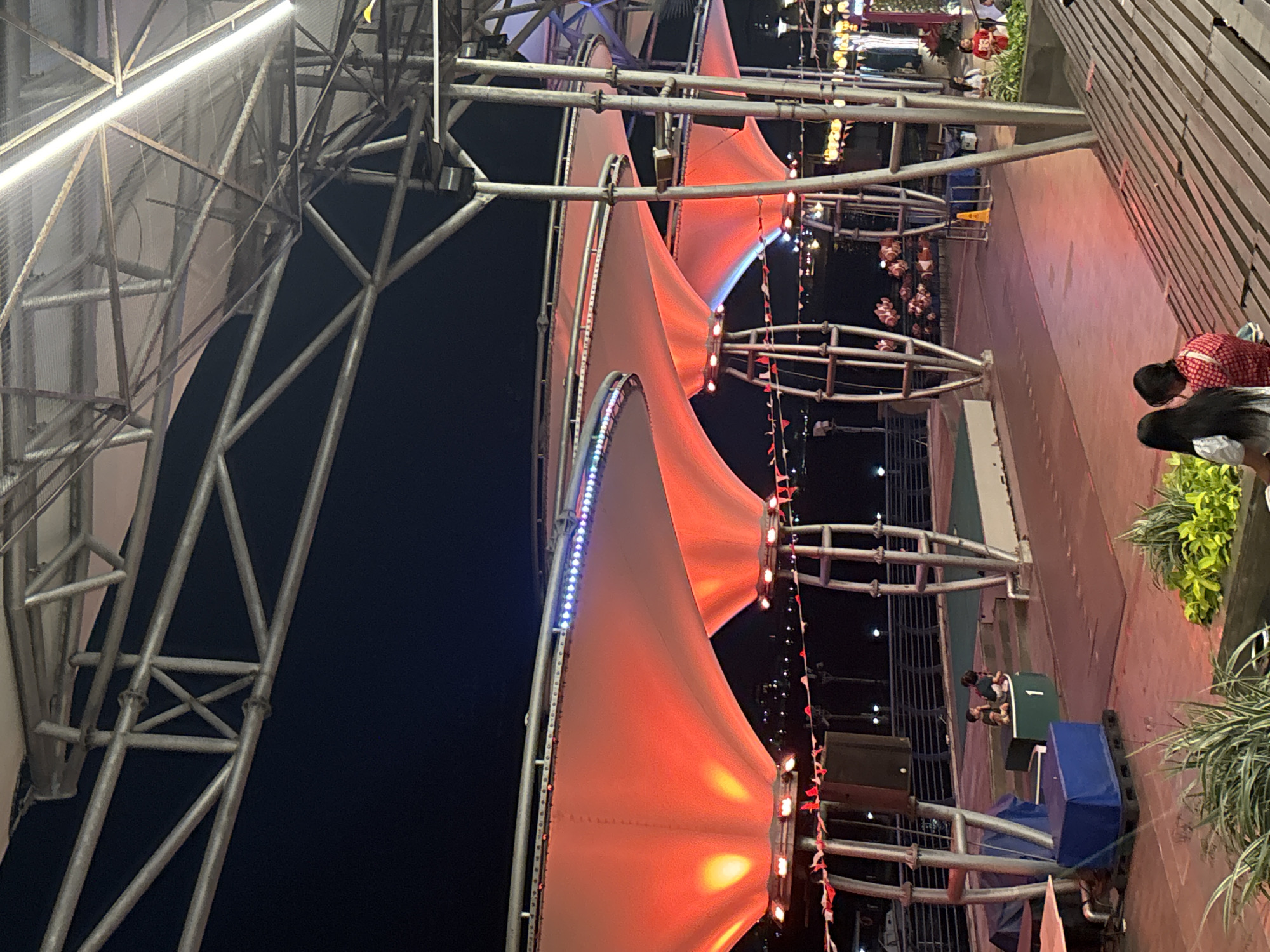 An outdoor evening scene in North Jakarta, Indonesia shows a waterfront promenade with a large metal and fabric canopy structure. The canopy, illuminated with red lights from below and string lights along its upper edge, is supported by tall metal poles. Below the canopy, a pathway runs alongside a railing overlooking a body of water. Several people are visible on the promenade, some sitting on tiered wooden seating in the foreground and others near a green platform with the number "1" on it. The background features the city's skyline with numerous lights, suggesting it is nighttime. The atmosphere appears to be a public gathering space, possibly a park or entertainment area.