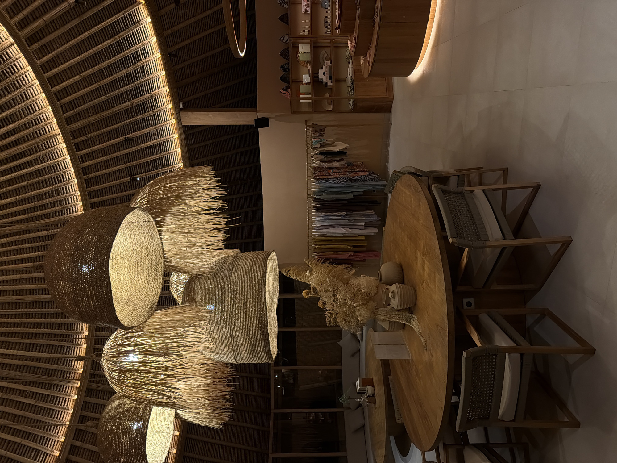 The image displays the interior of a commercial establishment in Unknown, Indonesia. A high, domed ceiling constructed from woven natural materials, possibly thatch or bamboo, dominates the upper portion, featuring exposed support beams and a continuous warm light strip along its base. Multiple large pendant light fixtures, made of woven natural fibers and some with long, frayed elements, hang from the ceiling, emitting a warm, amber glow.
In the mid-ground, two large, circular wooden tables are present. The table in the foreground is surrounded by chairs with woven backs and light-colored cushioned seats. On this table, a prominent arrangement of dried botanical elements, including grasses and flowers, is displayed in various neutral-toned ceramic vessels. Behind this, to the left, a second similar round wooden table is positioned in front of a long, curved, light-grey upholstered sofa. Large glass window panels behind the sofa reveal a dark exterior, indicating night conditions.
The right side of the room is dedicated to a retail area. A metal clothing rack holds various garments in muted colors. Adjacent to this, two wooden shelving units display an assortment of packaged goods, bottles, and decorative items. A distinct, curved, tiered wooden display counter with an illuminated base is located on the far right. The floor consists of large, light-colored square tiles. The walls are plain and light-toned. There are no individuals present in the scene, and no actions or interactions are depicted. The environment is uniformly illuminated by artificial warm lighting, highlighting the natural material aesthetic.