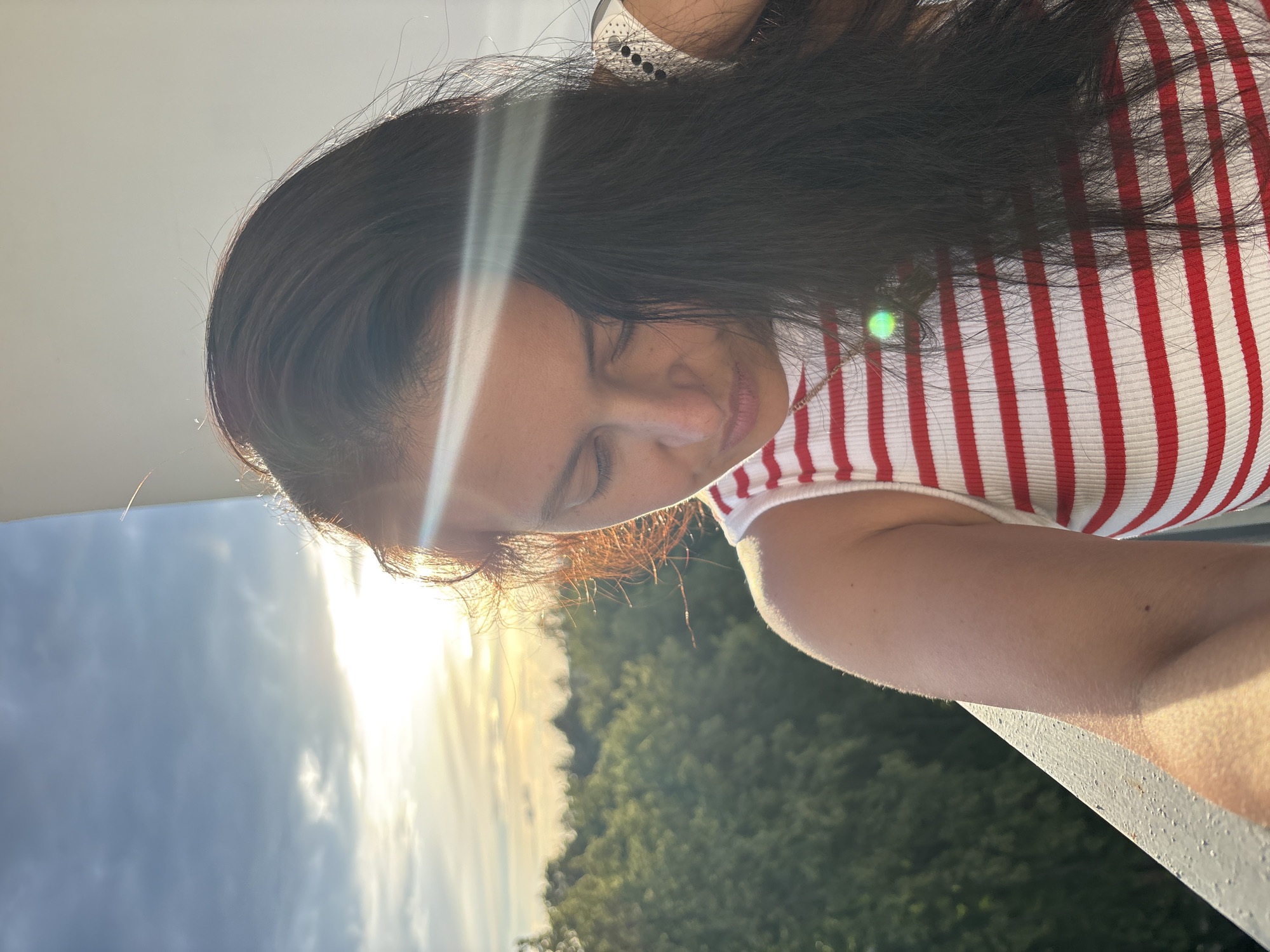 The media file shows a close-up, selfie-style shot of a person with dark hair and closed eyes, wearing a white and red striped top. The person's face is tilted downwards and bathed in soft, warm sunlight. A lens flare streaks across the image from the left. The background consists of a blurry, green landscape of trees and a sky with dramatic clouds, suggesting it is either early morning or late afternoon. The light and the person's relaxed expression evoke a sense of peace and tranquility. The image appears to be taken outdoors, possibly on a balcony or elevated vantage point given the presence of a railing in the foreground. There is no visible text in the image.