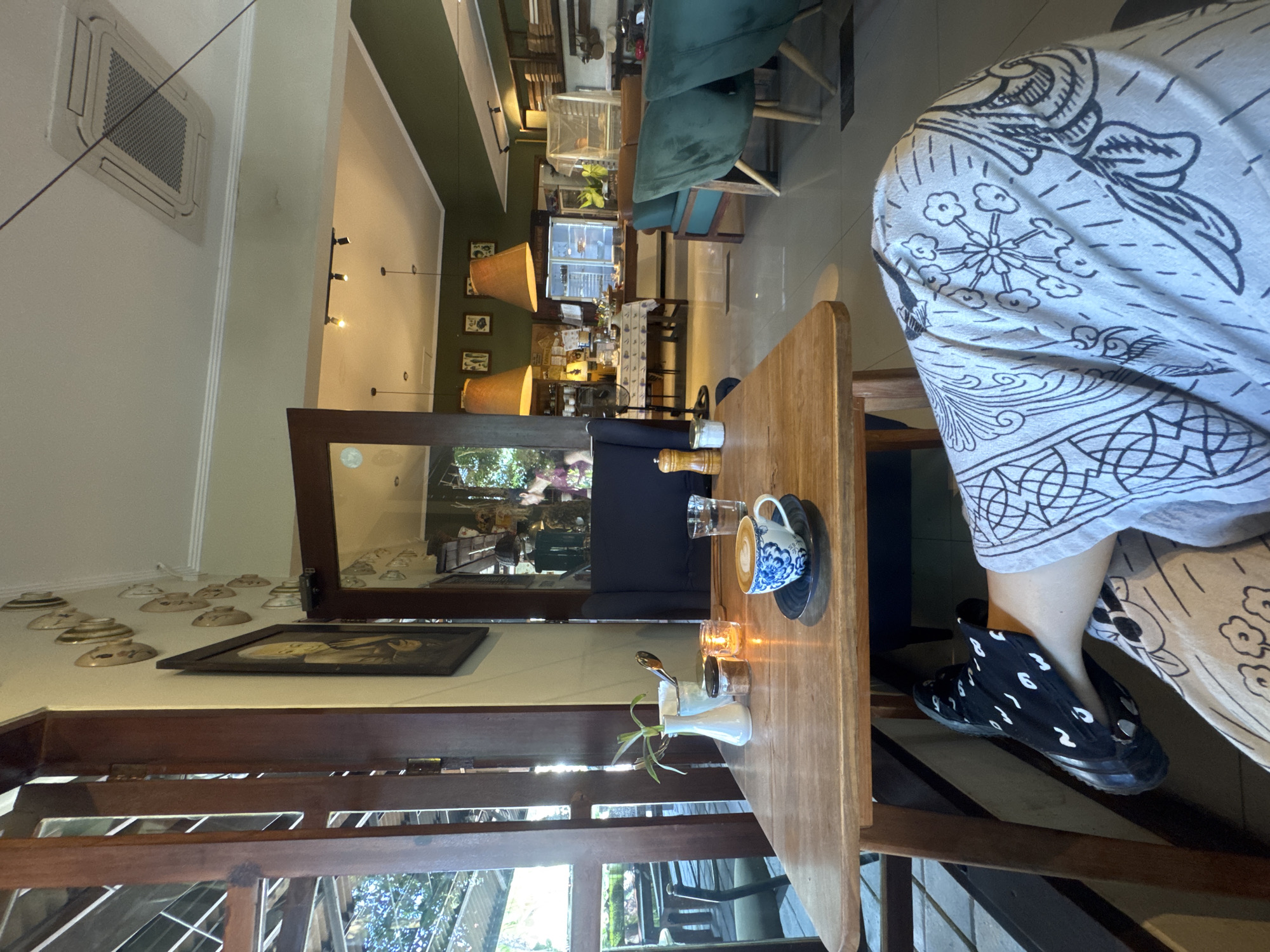 The image shows an interior shot of a cafe, possibly in North Kuta, Indonesia. The scene features a wooden table with a cup of coffee, a water glass, and a small vase with greenery. A person wearing a printed shirt and black patterned shoes is seated at the table. Outside the window, the view shows greenery and possibly urban structures. Inside the cafe, there are decorative plates and framed artwork along the walls. The space extends into a larger area with green chairs and other tables, suggesting a restaurant or cafe setting. Overhead, there are pendant lights and a view of the ceiling, which includes an air conditioning unit.