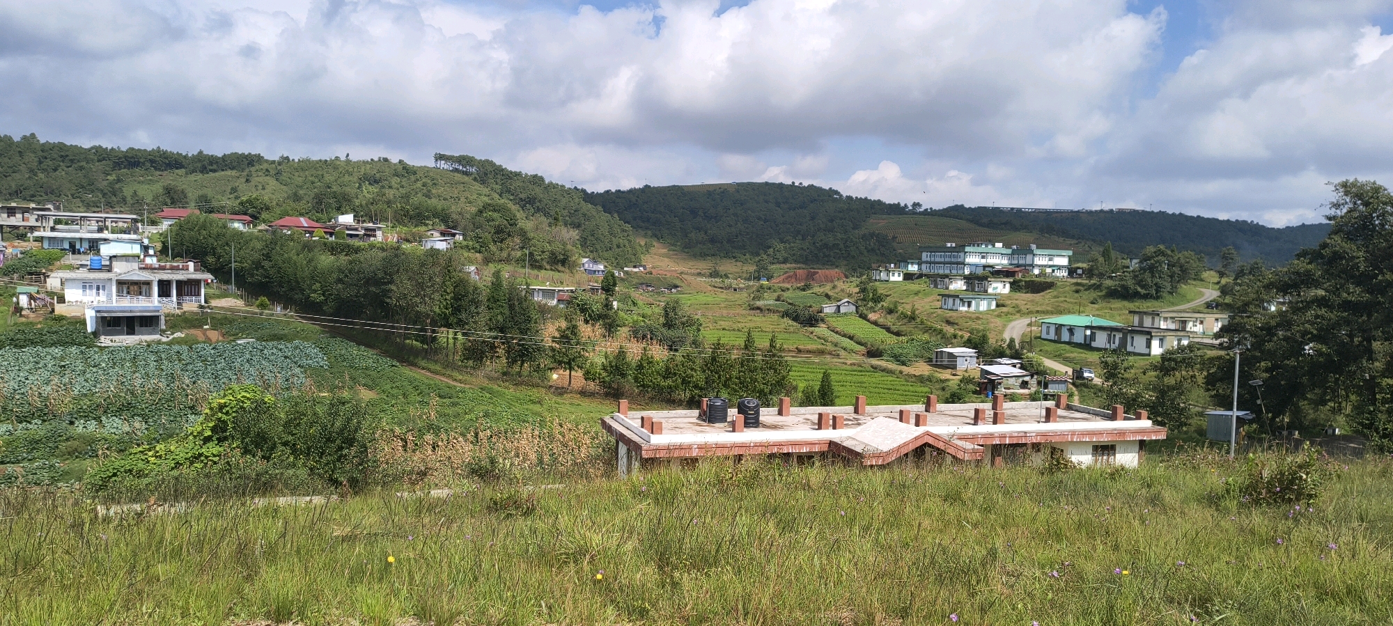 This is a wide, eye-level, outdoor, daytime photograph of a rural or semi-rural community nestled in rolling hills. The scene is dominated by green vegetation, including cultivated fields and trees, set against a backdrop of lush, forested hills. Several buildings are visible, varying in size and style. In the foreground, a low-rise building with a brick parapet and a flat roof is prominent. Further back, clusters of houses and larger institutional buildings, likely educational or administrative, are scattered across the landscape. The sky is partly cloudy with white cumulus clouds. A dirt road winds through the settlement. The overall impression is one of a tranquil, developing area with agricultural activity integrated into the built environment. This scene is located in Sohiong, India.