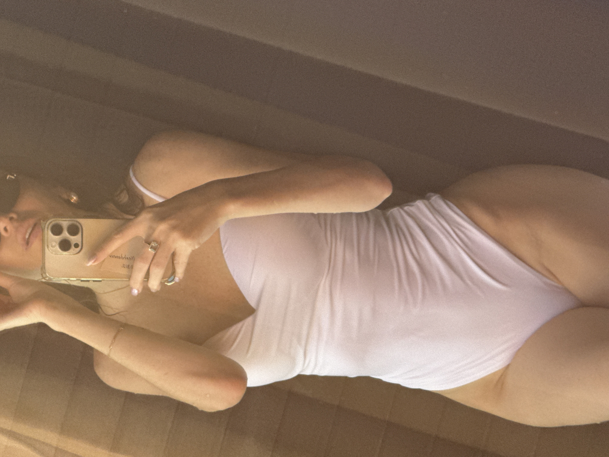 The image shows a woman in a white one-piece swimsuit taking a mirror selfie. She is holding a gold-colored smartphone, which has a distinct camera bump. She is wearing a ring on her right hand. The background appears to be a textured wall, possibly made of wood or a similar material, with vertical lines. The lighting suggests an indoor setting, possibly a bathroom or dressing room. There is no indication of a specific location in Dubai, United Arab Emirates.