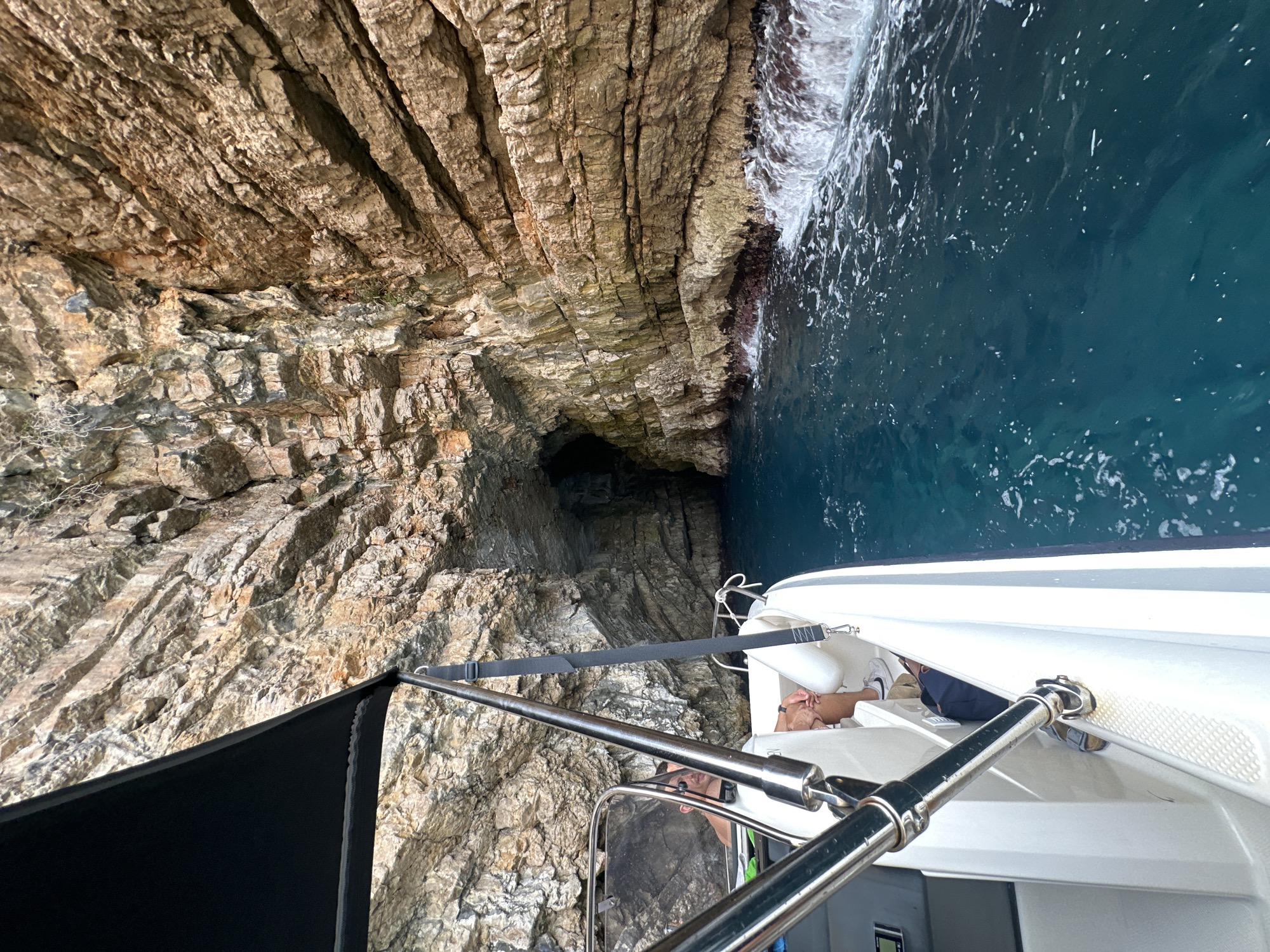 A boat approaches a cave entrance in a rocky cliff face on the Luštica peninsula, Montenegro. The boat is white with a black canopy and chrome railings. Inside the boat, two individuals are visible. The driver, a man with sunglasses and a partially visible torso, is at the helm. Another person, whose legs are visible, is seated. The water is a deep blue, with waves crashing against the base of the cliff. The cliff face is layered and textured, with vegetation growing in some crevices. The cave entrance is dark and partially submerged.