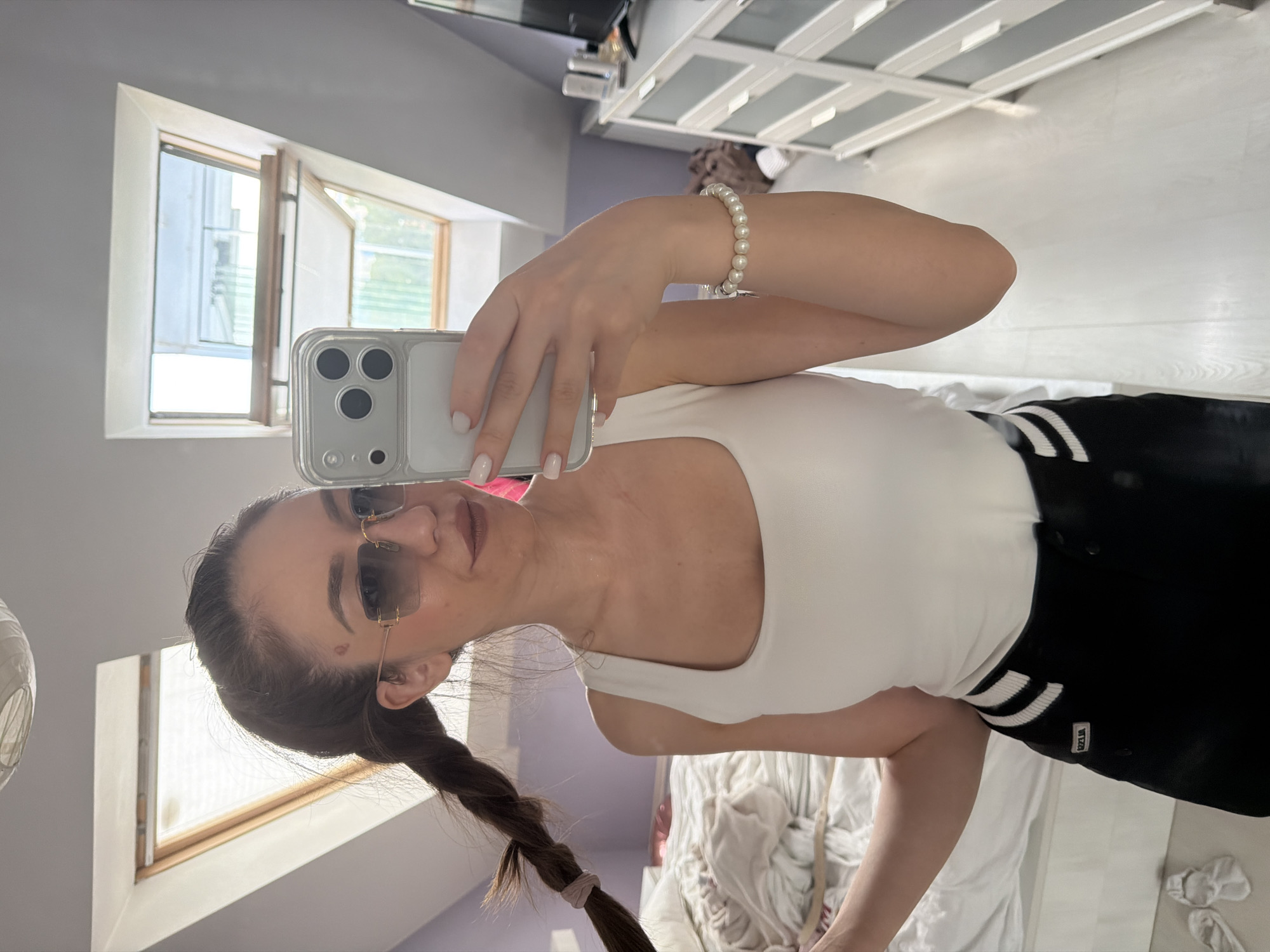 A mirror selfie captures a young adult female in a domestic bedroom setting within the city of Sofia, Bulgaria. The female subject has light skin, dark brown hair styled in a single high braid secured with a light pink elastic, and wears light brown lipstick. She is adorned with rimless rectangular sunglasses featuring gradient gray lenses and gold-toned frames, a plain white square-neck tank top, and black shorts/trousers with two white horizontal stripes on the side. A small white label with "EZZUA" is visible on her left hip. A white pearl bracelet is present on her right wrist, and her fingernails are painted white. A small reddish mark is visible on her forehead slightly above the bridge of her nose.
The subject's right hand holds a light-colored smartphone (identified as an iPhone due to the triple camera array) encased in a clear protective cover, reflecting her image. The background reveals a room with light purple walls on the left and light gray walls on the right, indicative of an attic or top-floor space due to a slanted ceiling. Two open skylight windows are visible in the ceiling, showcasing bright outdoor conditions and green foliage. To the immediate left, an unmade bed with white bedding and a pair of white slippers on the floor are partially visible. To the right, a white multi-drawer dresser with silver handles stands against the wall, topped with various objects and a black flat-screen television set. The flooring is a light-colored material, possibly wood or laminate. An overhead ceiling light fixture is partially visible at the top center.