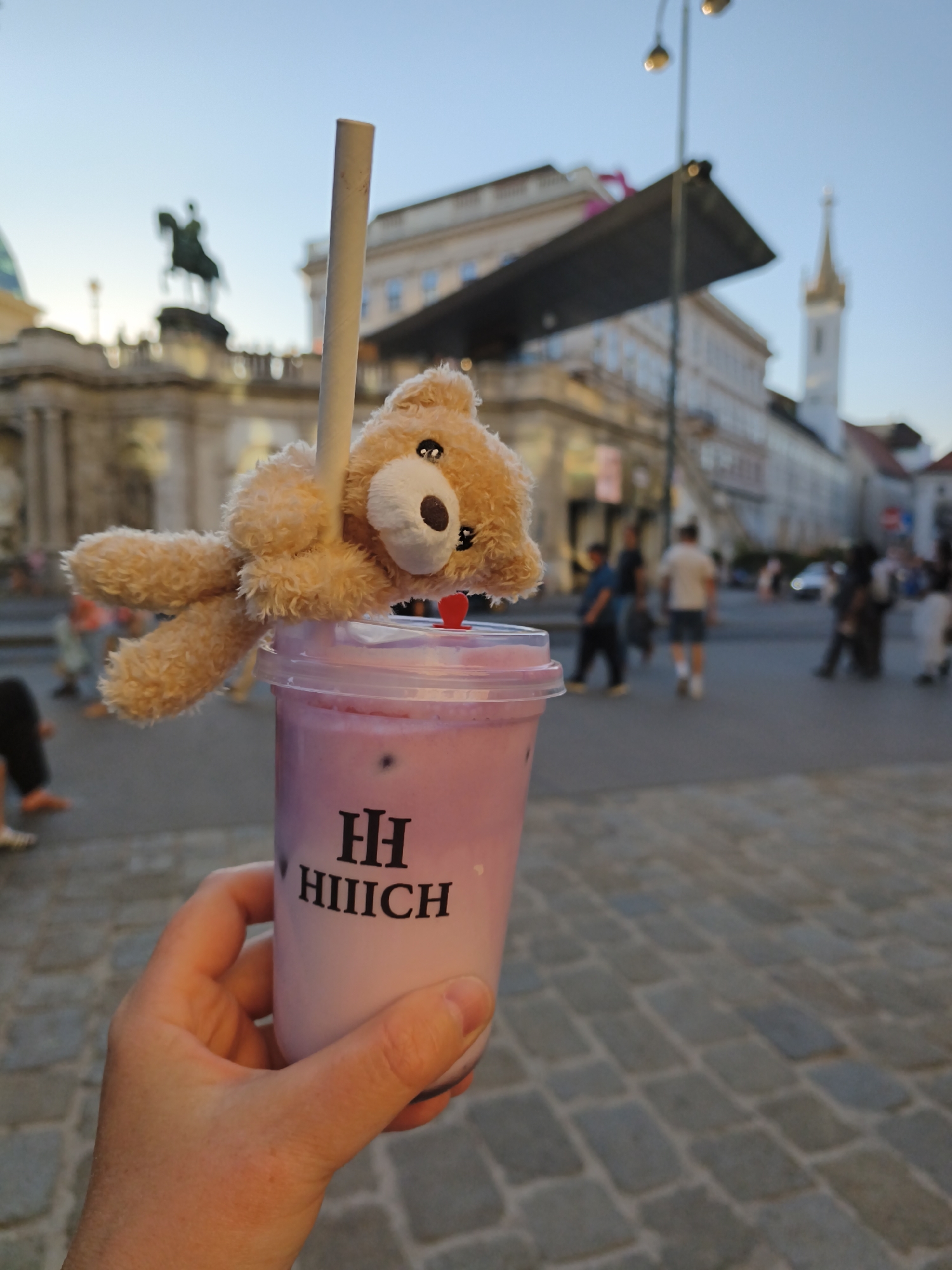 A close-up shot shows a hand holding a pink beverage in a plastic cup. A small stuffed teddy bear is draped over the straw, which is inserted into the cup. The cup has a black logo that reads "HH HIIICH." In the background, people are walking on a cobblestone street. Buildings and a church spire are visible, suggesting an urban setting. The lighting indicates it is either early morning or late afternoon. This scene appears to be in Vienna, Austria, near St. Stephen's Cathedral, given the architectural style and presence of a church spire common in the city.