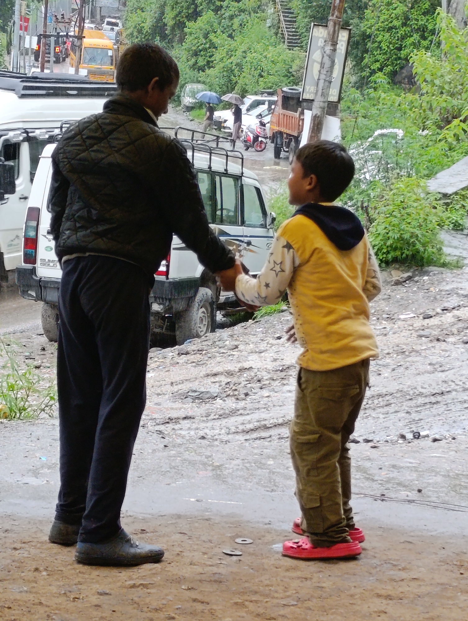 In Aleo, India, two individuals are standing on a wet, unpaved road. The person on the left, appearing to be an adult male, is wearing a black quilted jacket and dark blue trousers. He is facing right and appears to be reaching out with his left hand, palm up. The person on the right, appearing to be a young boy, is wearing a yellow hooded sweatshirt with a black hood and dark green trousers. He is facing left and is holding out his left hand, palm down, towards the other individual. A white SUV-like vehicle is parked behind the adult male. In the background, there are trees, a yellow bus, and other vehicles, suggesting a street scene with moderate traffic and lush greenery. The ground is muddy and appears to have recently experienced rain.
